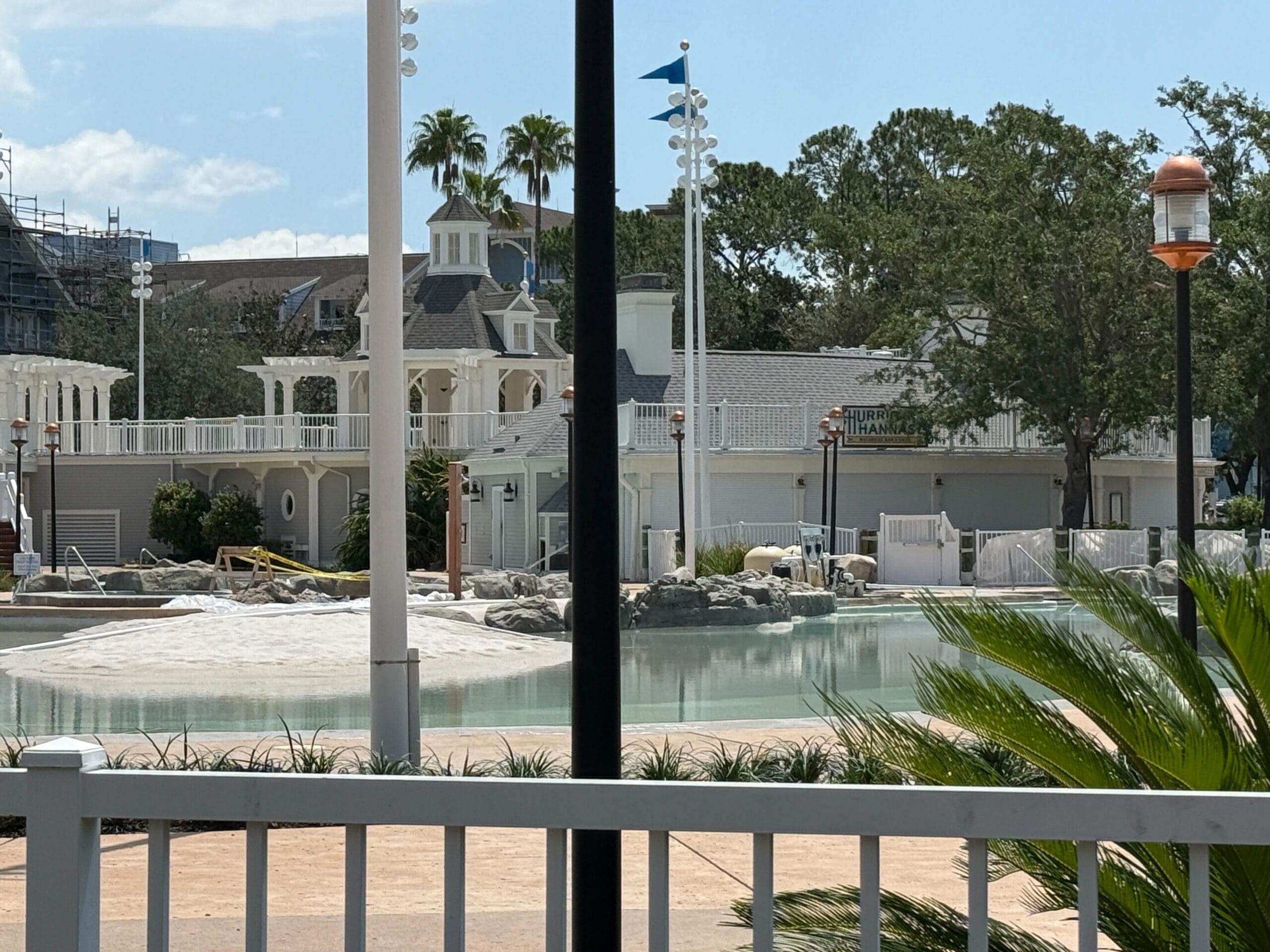 A resort pool area with calm water, rocks, and buildings, viewed through a white fence with palm fronds.