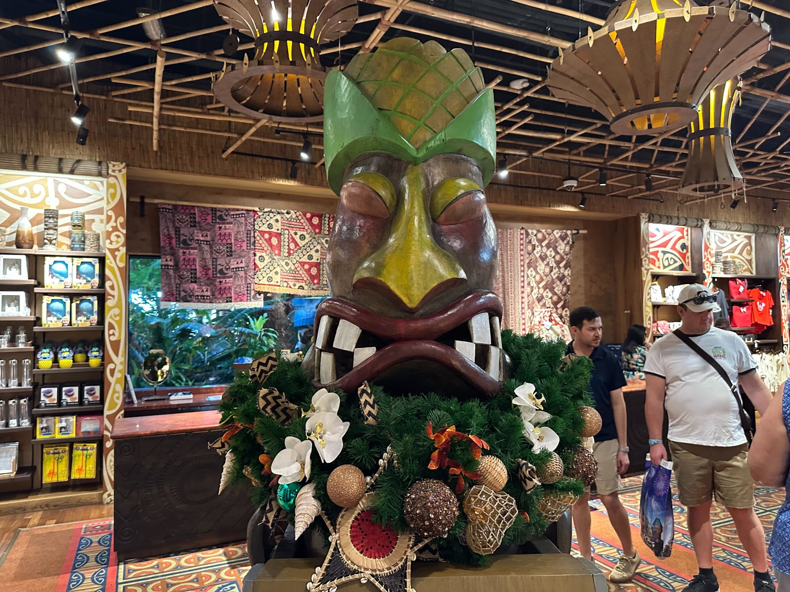 A colorful Tiki statue decorated with greenery and ornaments stands in a store. Shoppers and various merchandise are visible in the background.
