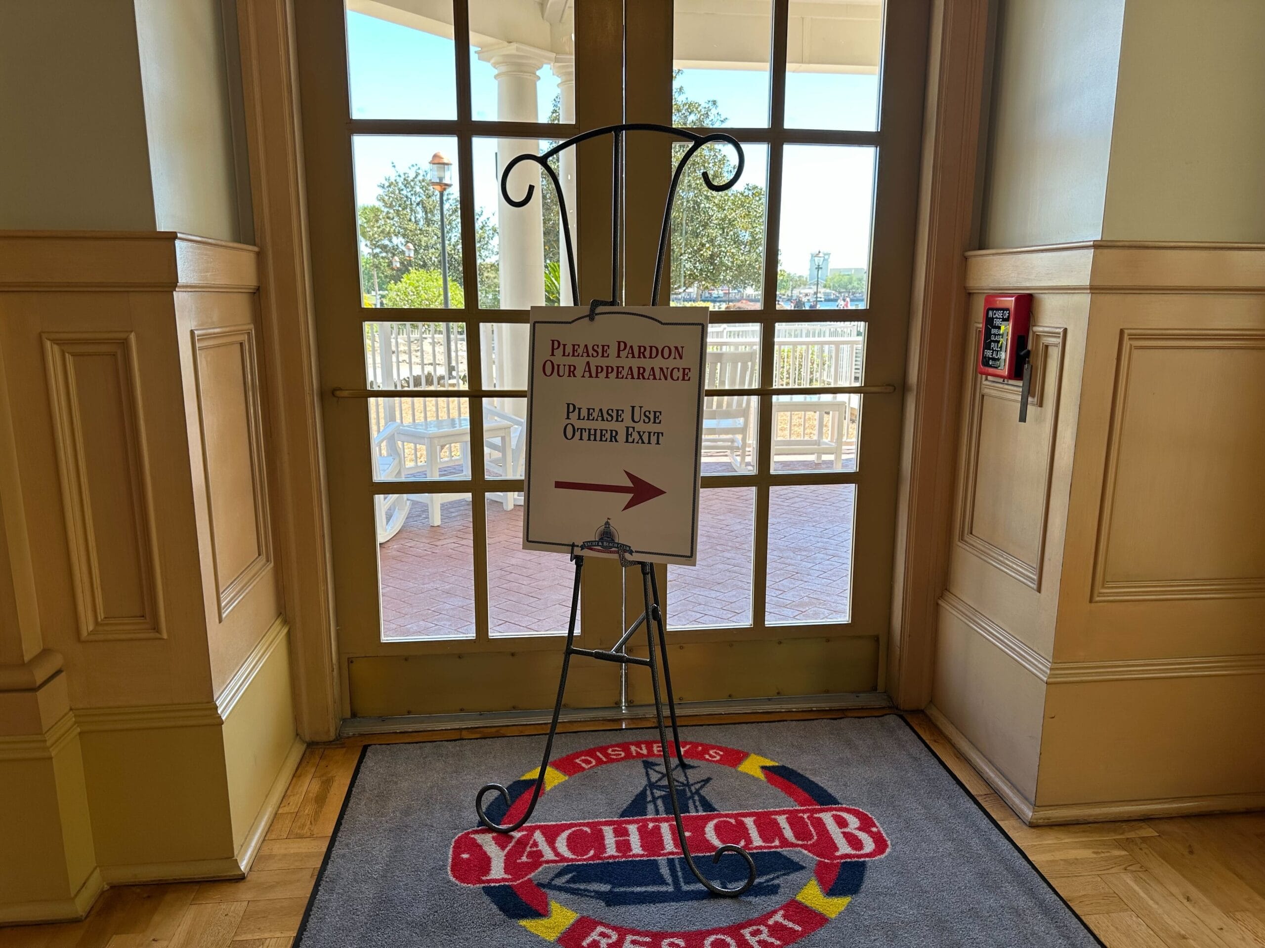 A sign on an easel at Disney's Yacht Club Resort directs guests to use an alternative exit due to ongoing construction. The backdrop features tile and a glimpse of the outdoor scenery, hinting at the nearby Stormalong Bay Pool.
