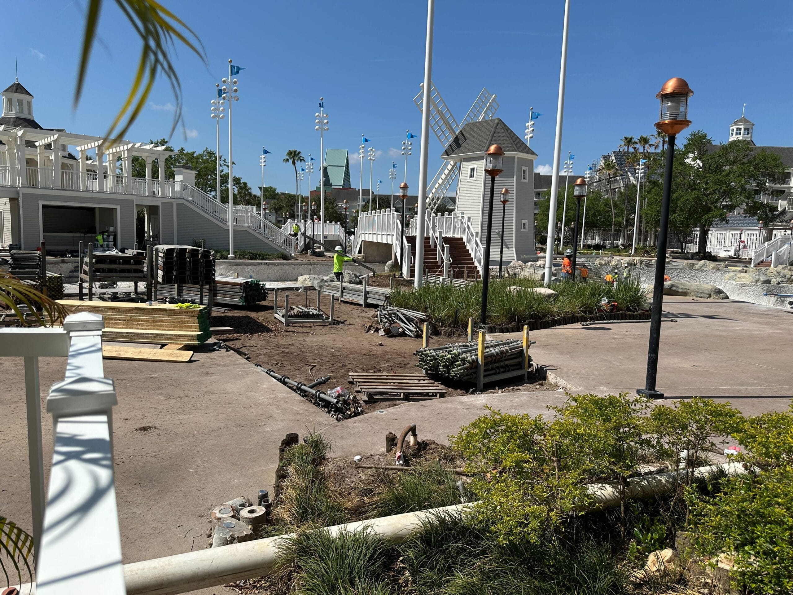 A construction site with a windmill structure stands tall, surrounded by workers and materials. The nearby greenery and buildings bask under a clear blue sky, evoking the tranquil charm of Disney's Yacht Club near Stormalong Bay.