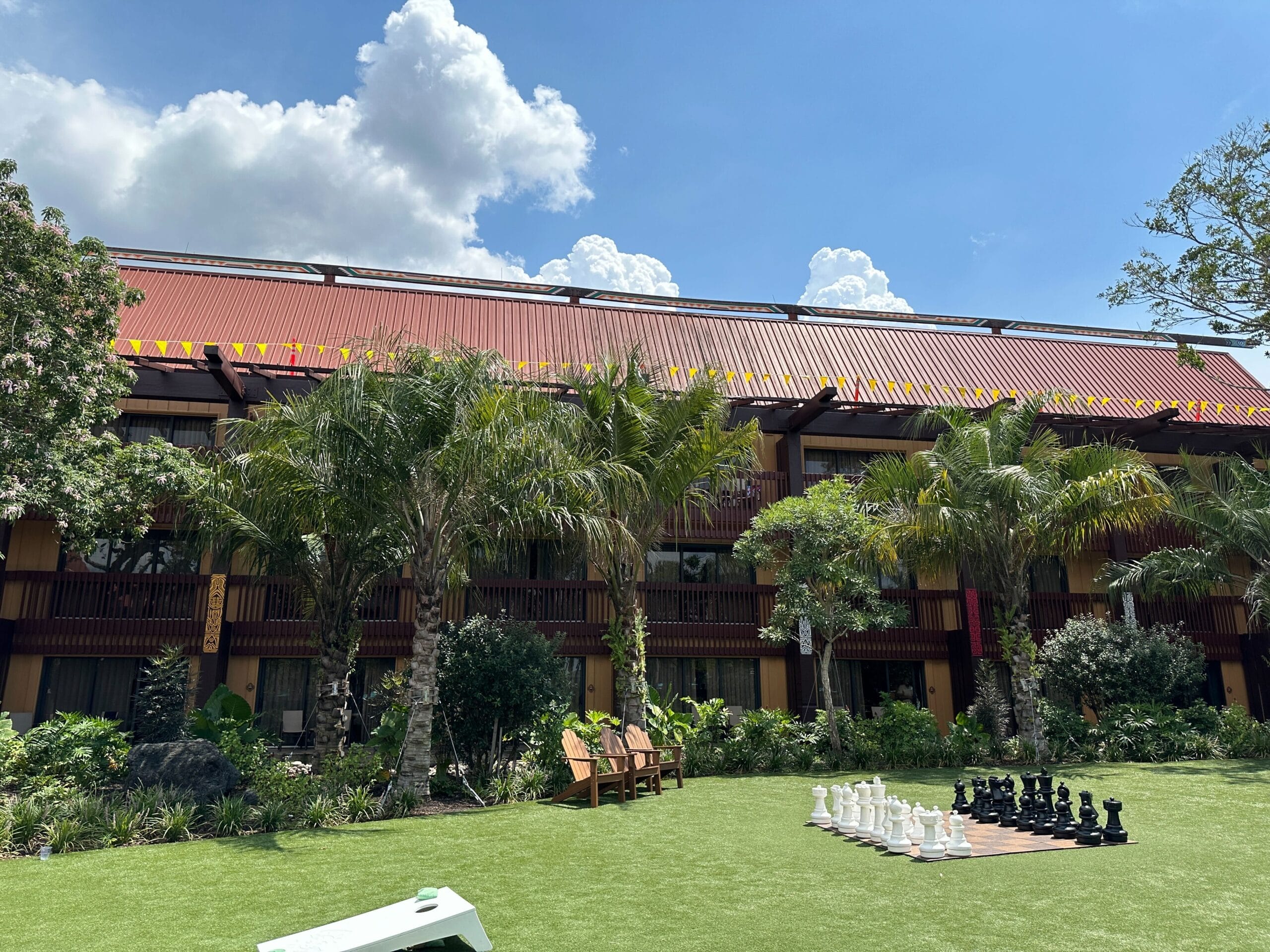A tropical resort courtyard with palm trees, benches, and a large outdoor chess set on green grass under a partly cloudy sky.