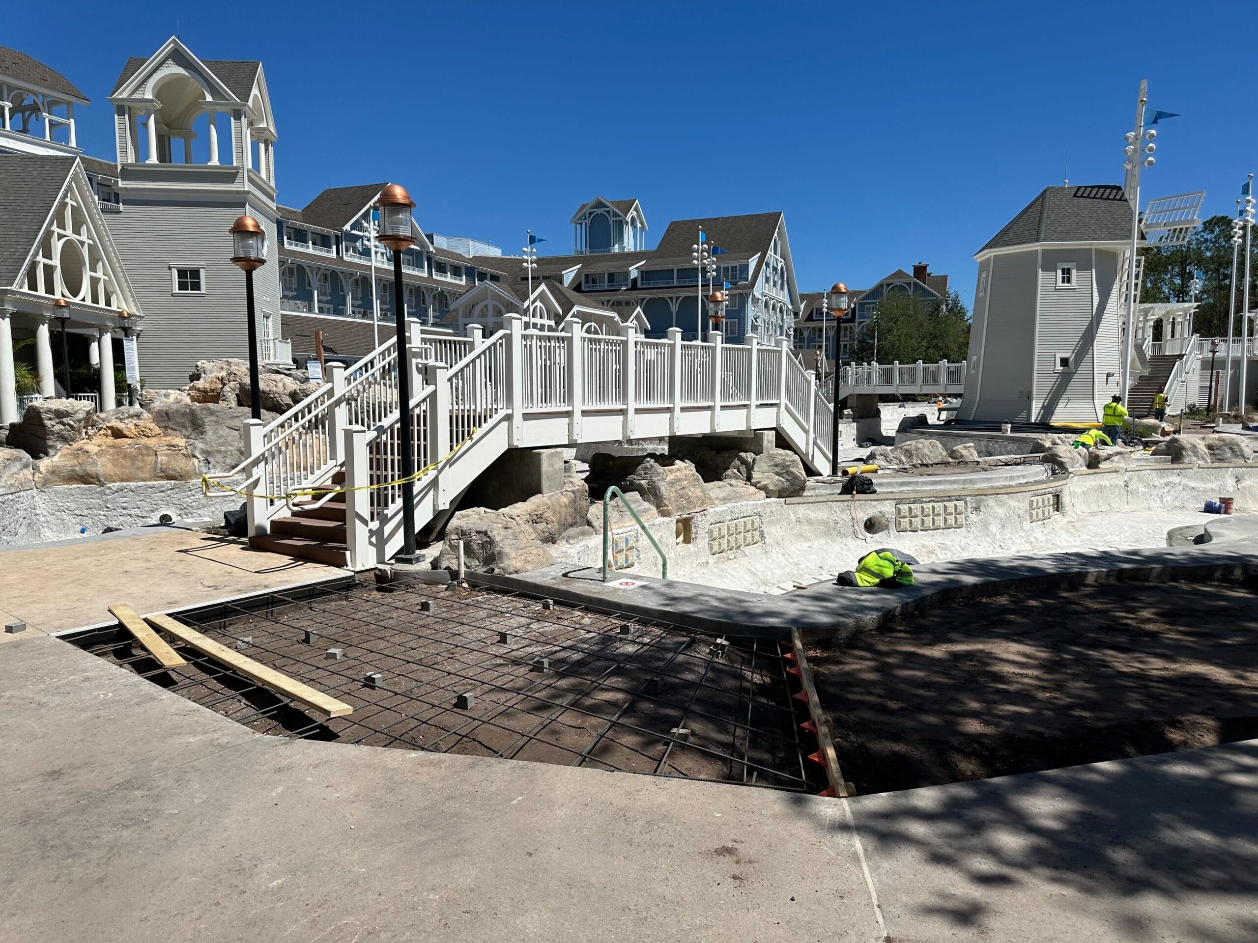 At Disney's Yacht Club, the construction site buzzes with activity near the Stormalong Bay Pool. Workers are busy near a bridge as buildings with gabled roofs stand gracefully in the background under a clear blue sky.