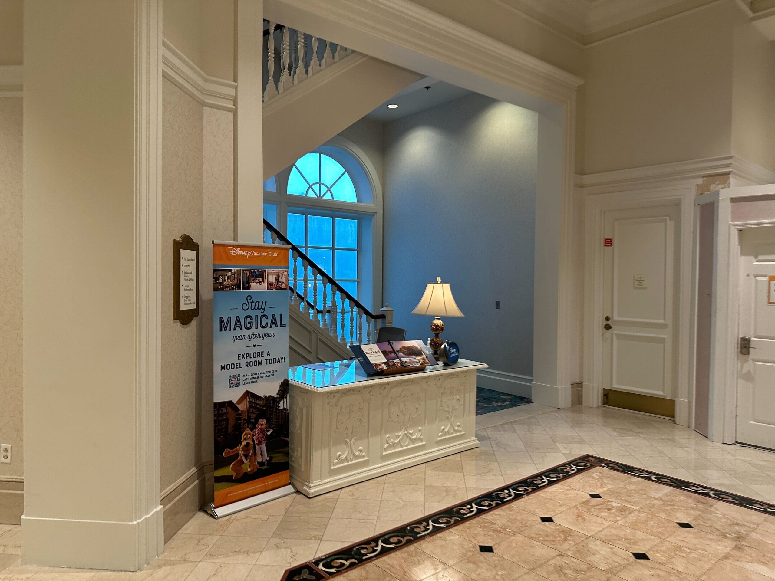 Hotel lobby with a white reception desk, lamp, Grand Floridian sign, and staircase in the background.