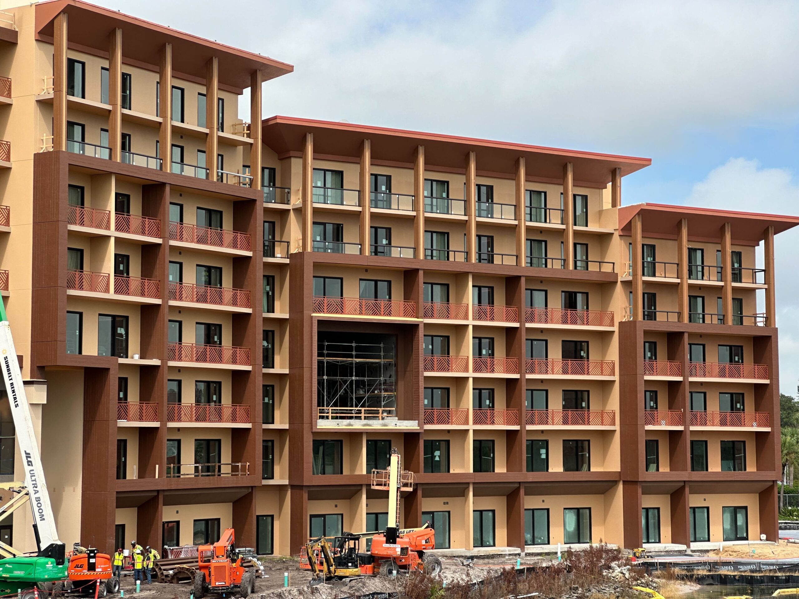 A multi-story building under construction with workers, construction equipment, and scaffolding visible. The exterior appears mostly complete with windows and balconies installed.