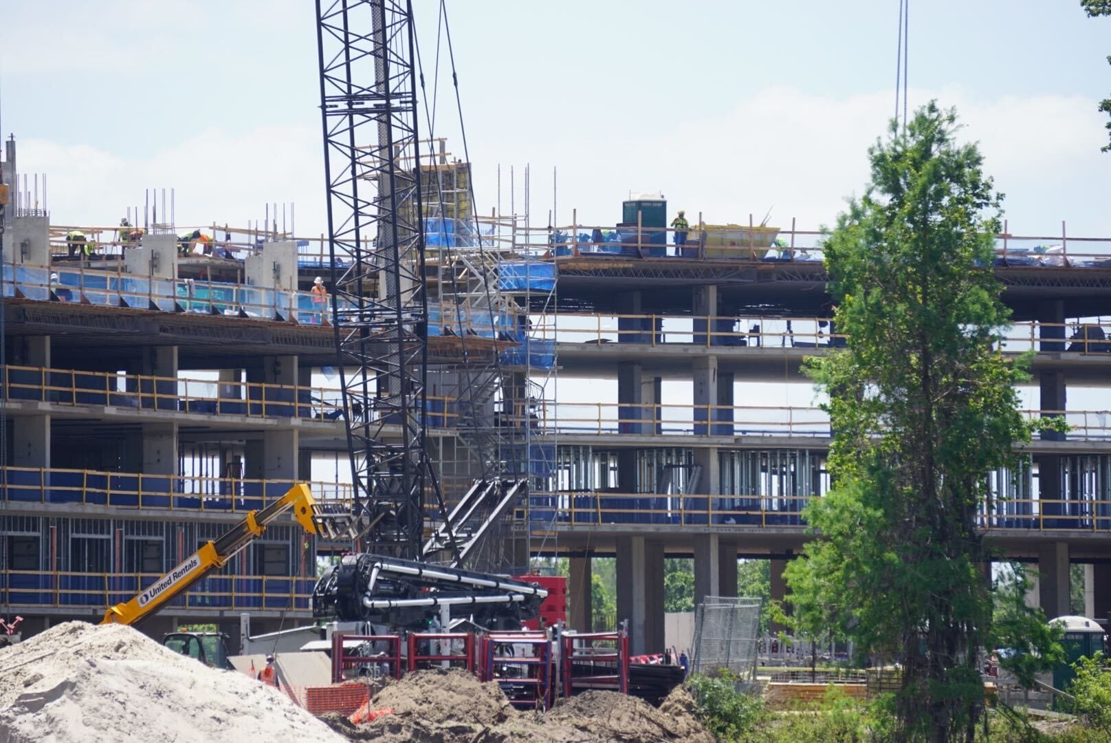 Construction site with a partially built multi-story building, cranes, and construction vehicles. Workers are visible on different levels. Trees are in the foreground. Sky is clear.