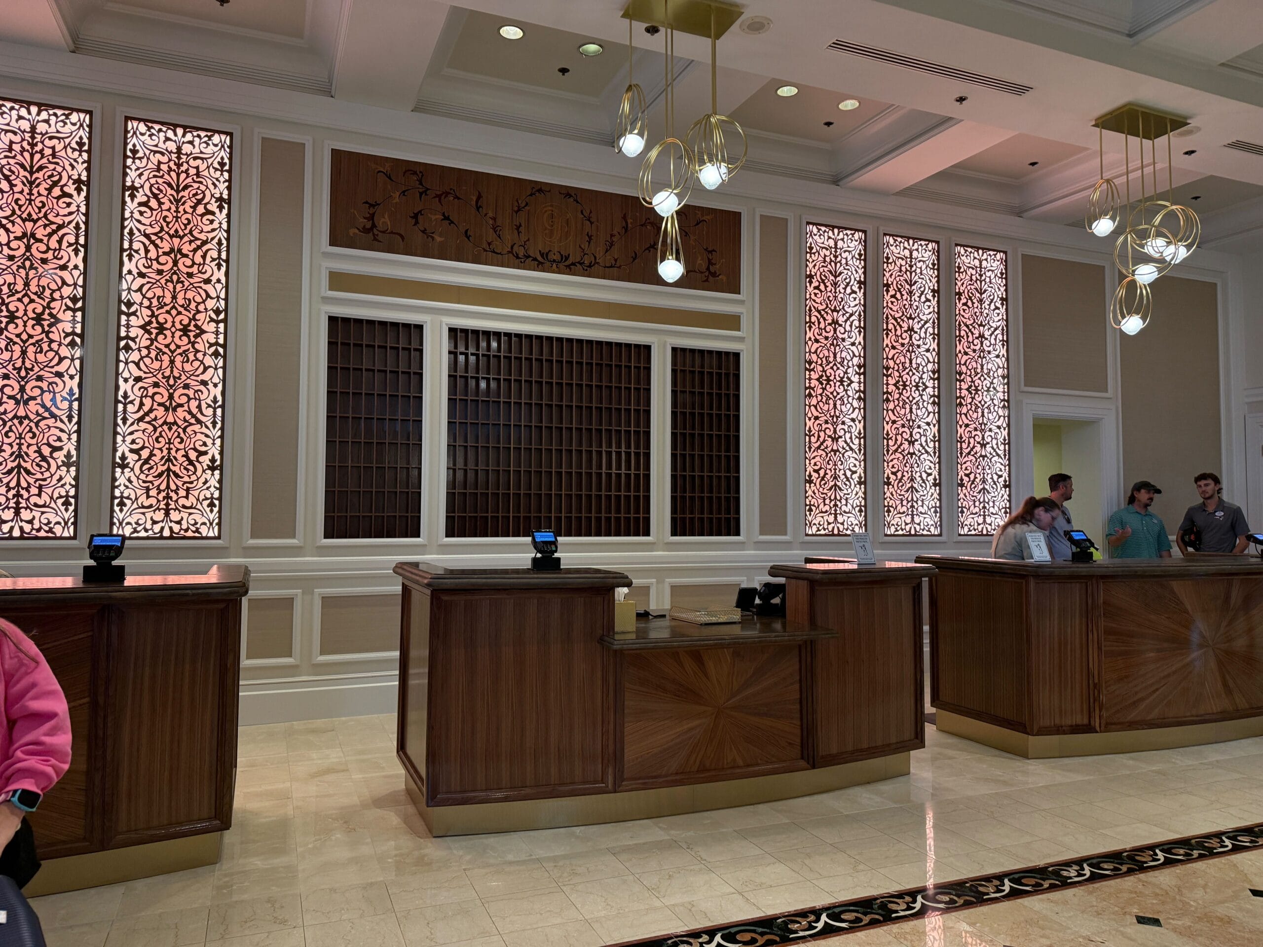 Hotel reception area with three wooden front desks, ornate light panels, and people checking in.