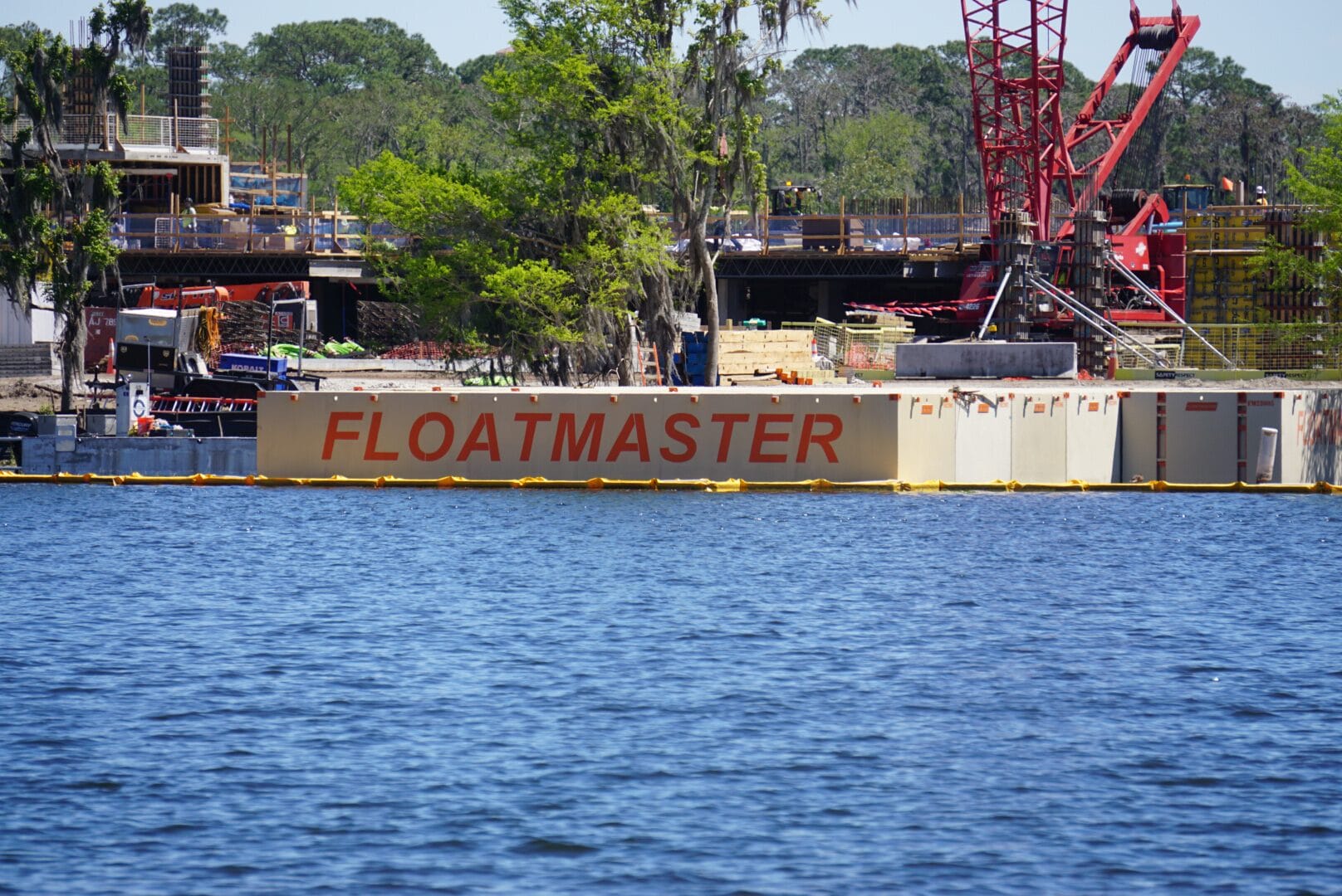 A floating platform labeled FLOATMASTER on a body of water, with construction equipment and trees in the background.