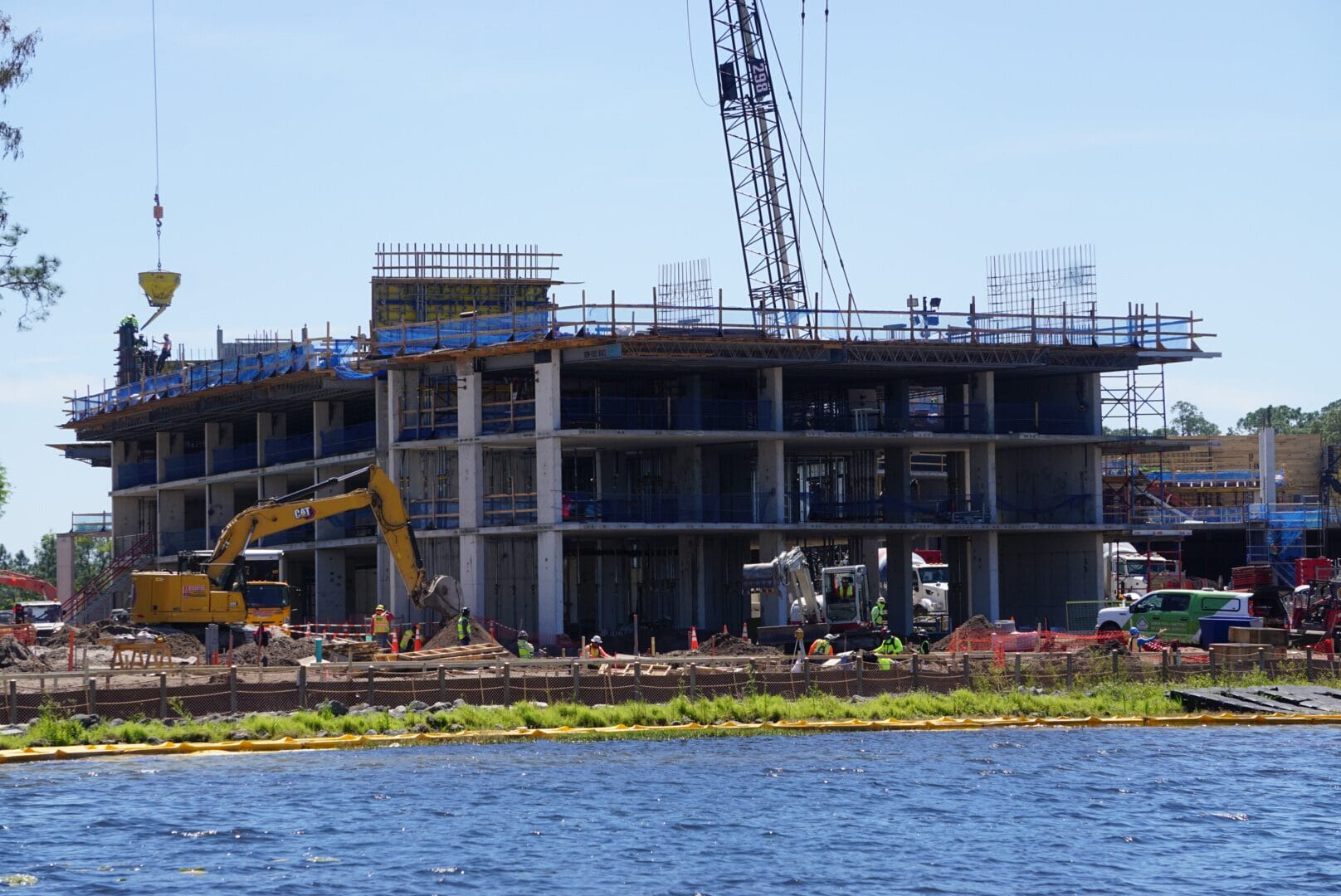 Construction site with a multi-story building under development, featuring cranes, scaffolding, and construction vehicles. Workers are present around the site, and water is visible in the foreground.