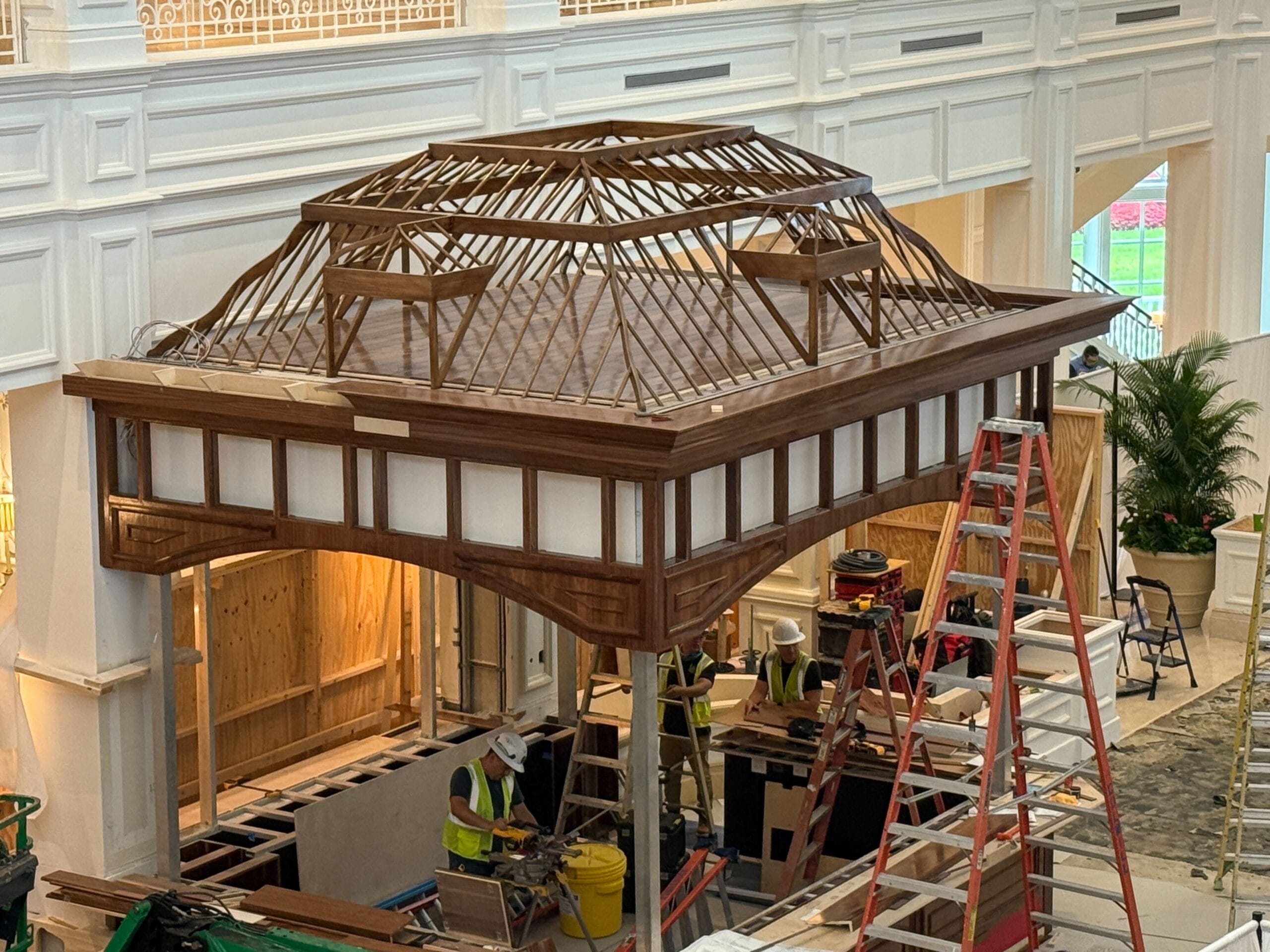 Workers construct a large wooden pavilion structure indoors, surrounded by ladders and building materials.