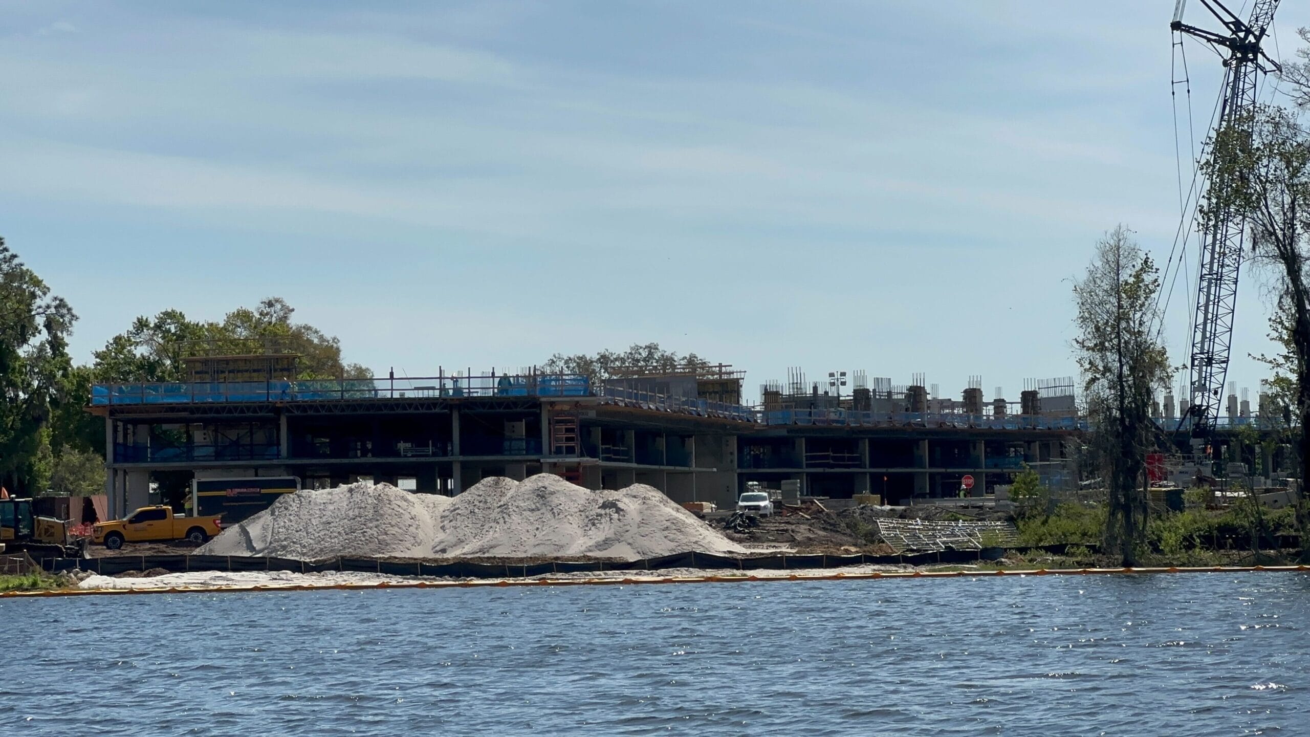 A partially built structure rises on the riverbank, surrounded by piles of sand and construction vehicles. A crane stands tall on the right under a clear sky, hinting at the transformation into a future Lakeshore Lodge.
