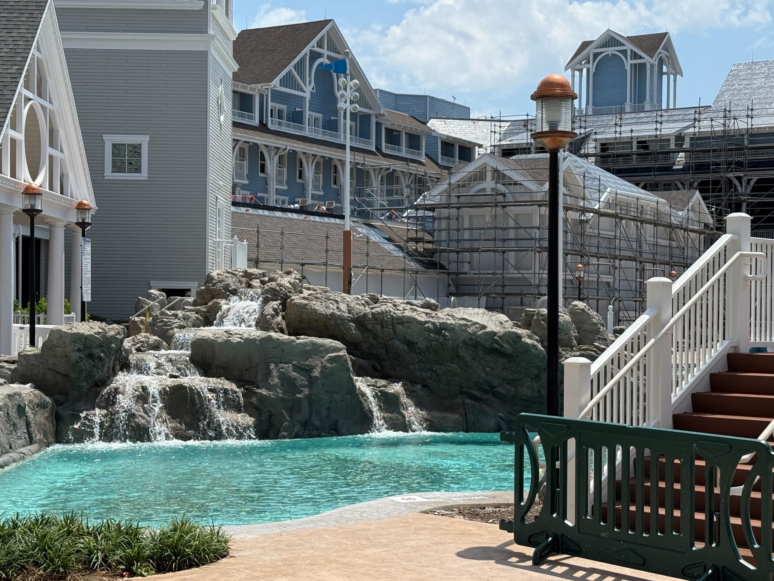 A pool with rock waterfalls, nearby construction, and a white staircase under a partly cloudy sky.