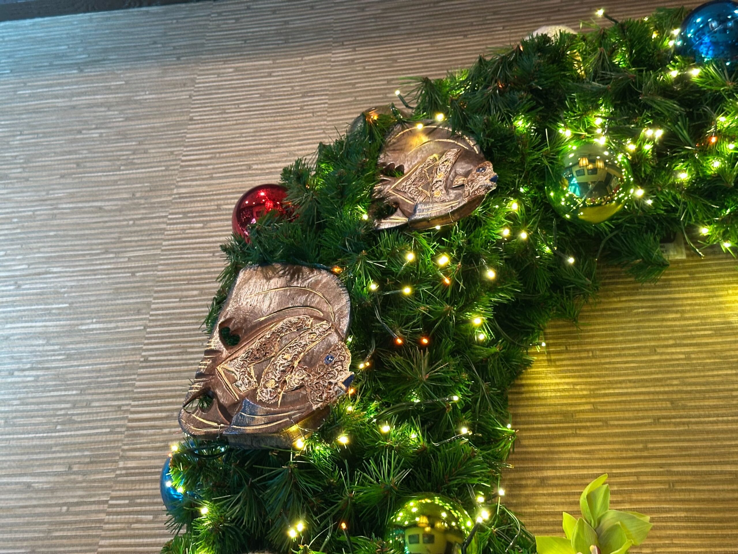 Close-up of a decorative wreath with greenery, colorful baubles, and metallic fish ornaments, adorned with small white lights on a textured beige wall.