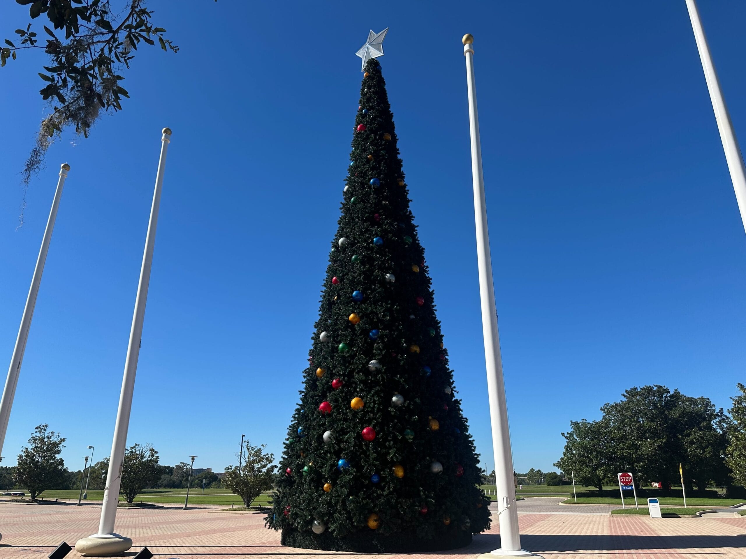 Tall Christmas tree with colorful ornaments and a star on top, surrounded by flagpoles, set against a clear blue sky.