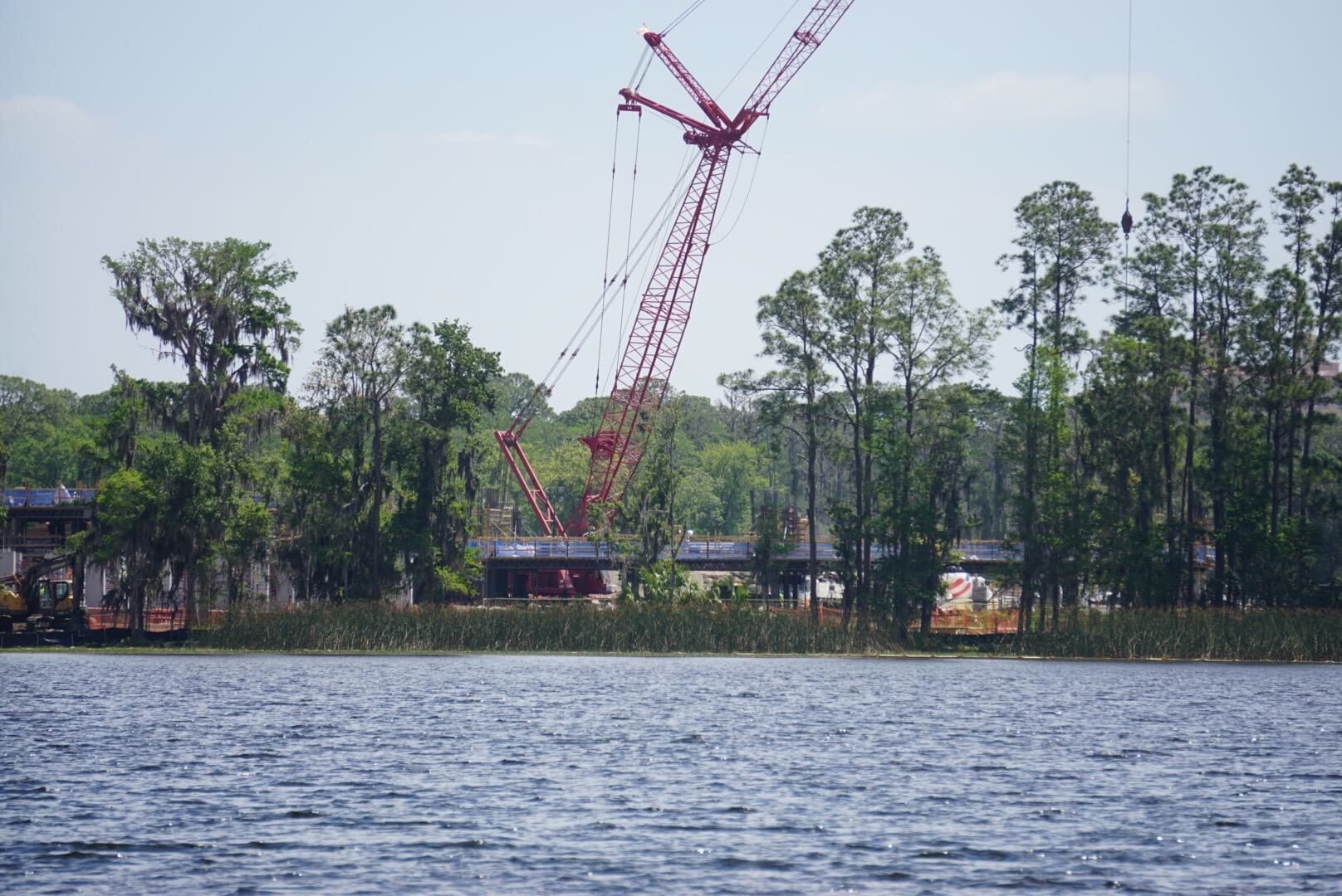 A large red crane operates near a lake, surrounded by tall trees and construction equipment.