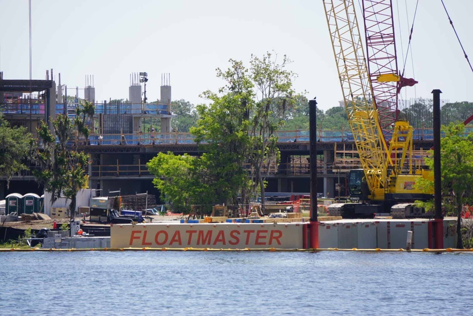 A barge labeled FLOATMASTER, sporting construction equipment like a yellow crane, glides across the water near the thrilling development site of Lakeshore Lodge. Stay tuned for more construction updates on this exciting project!.