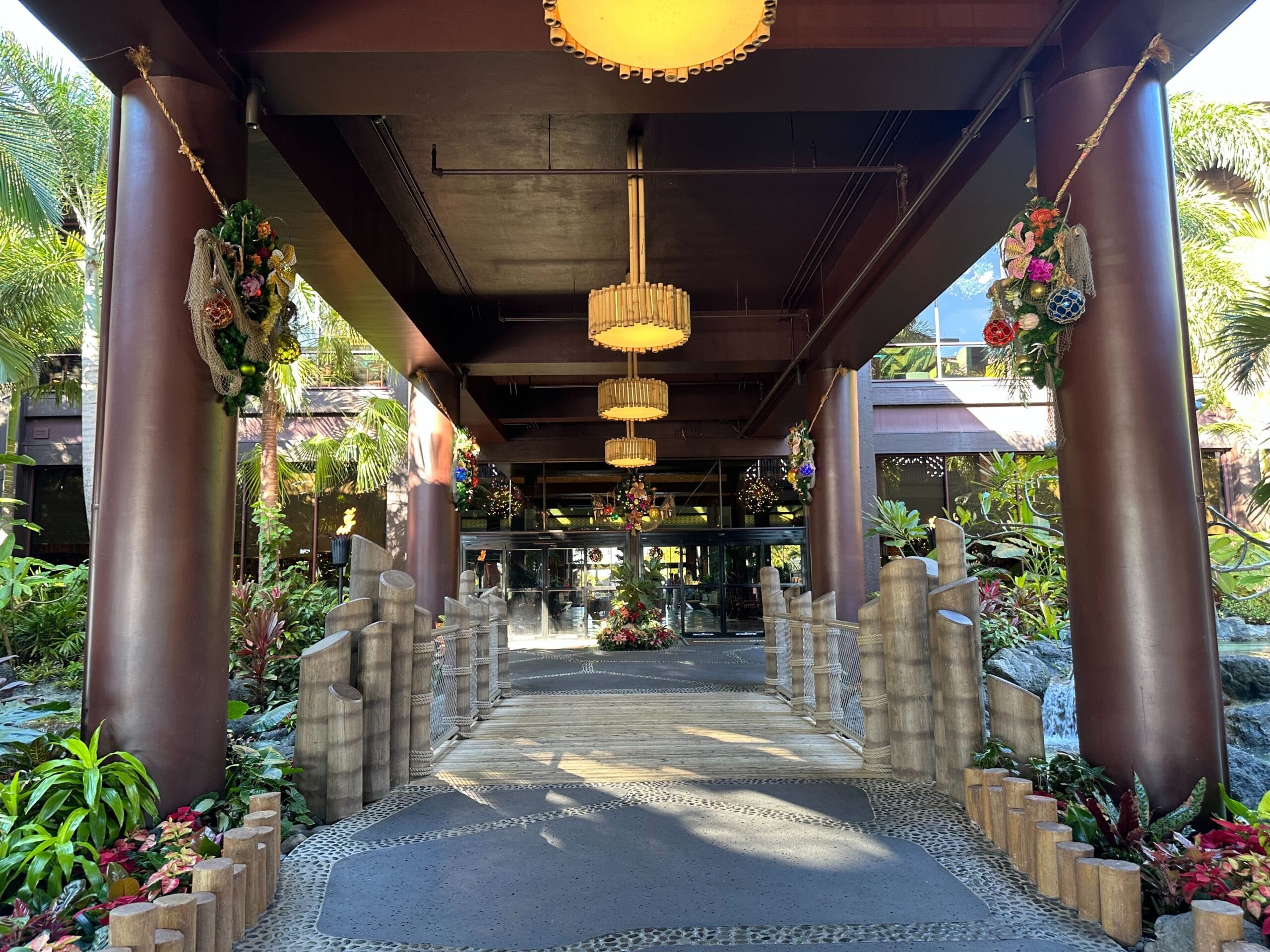 Covered walkway with hanging lights and floral decorations, surrounded by tropical plants and tall wooden pillars.