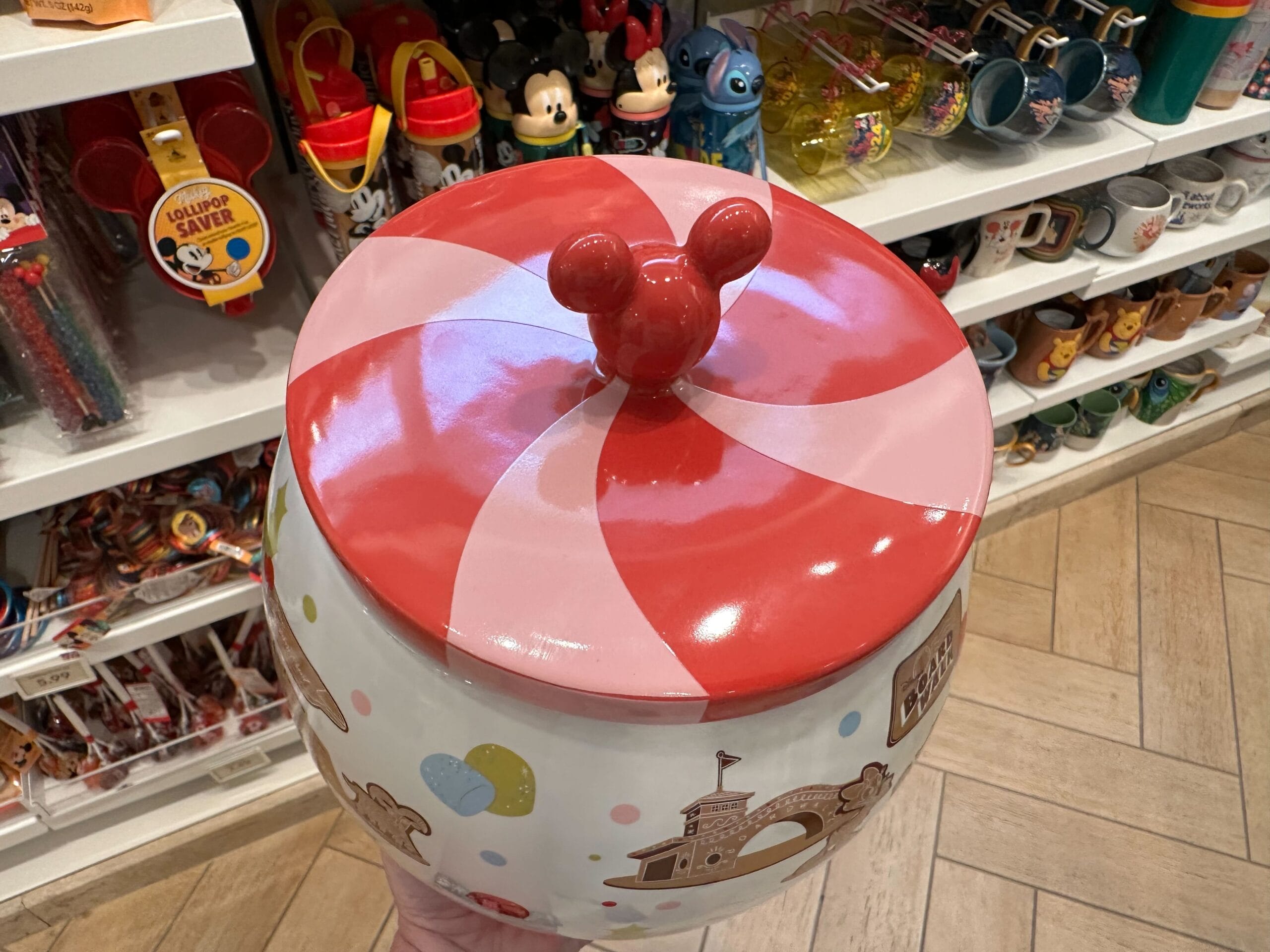 A hand holds a jar with a red and white swirl lid featuring mouse ears, surrounded by Disney-themed merchandise in a store reminiscent of the Grand Floridian Gingerbread House ambiance.