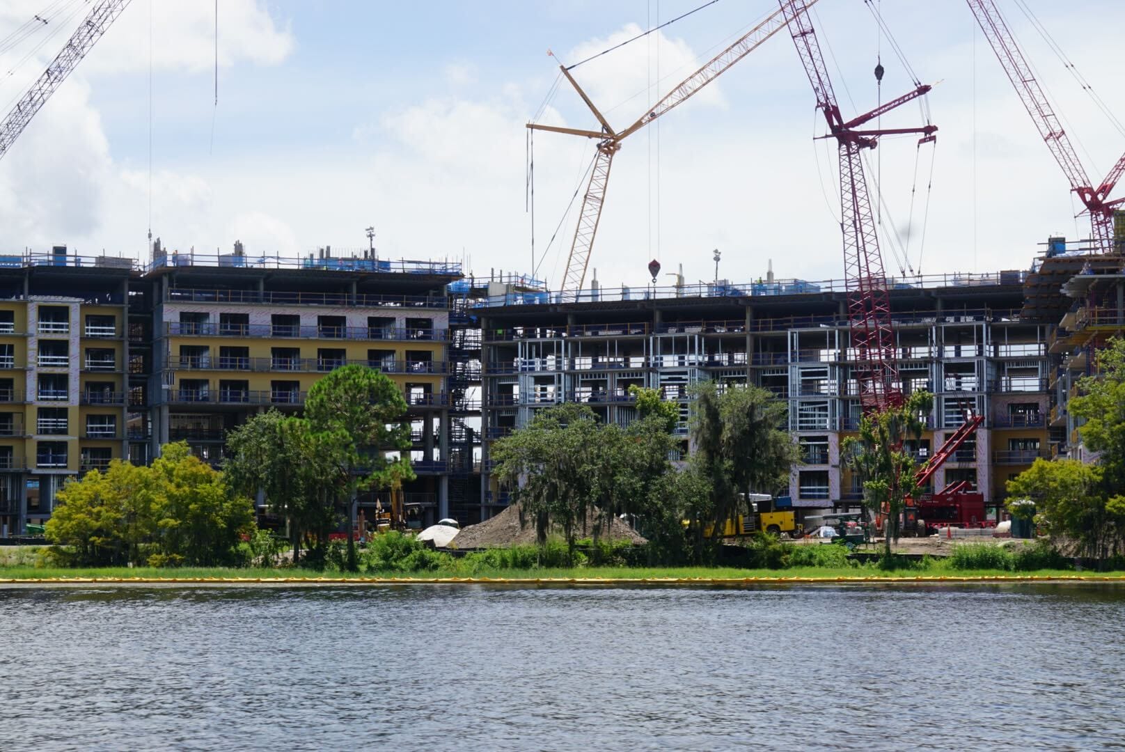 Construction cranes and equipment working on multi-story buildings near a body of water, with trees in the foreground.
