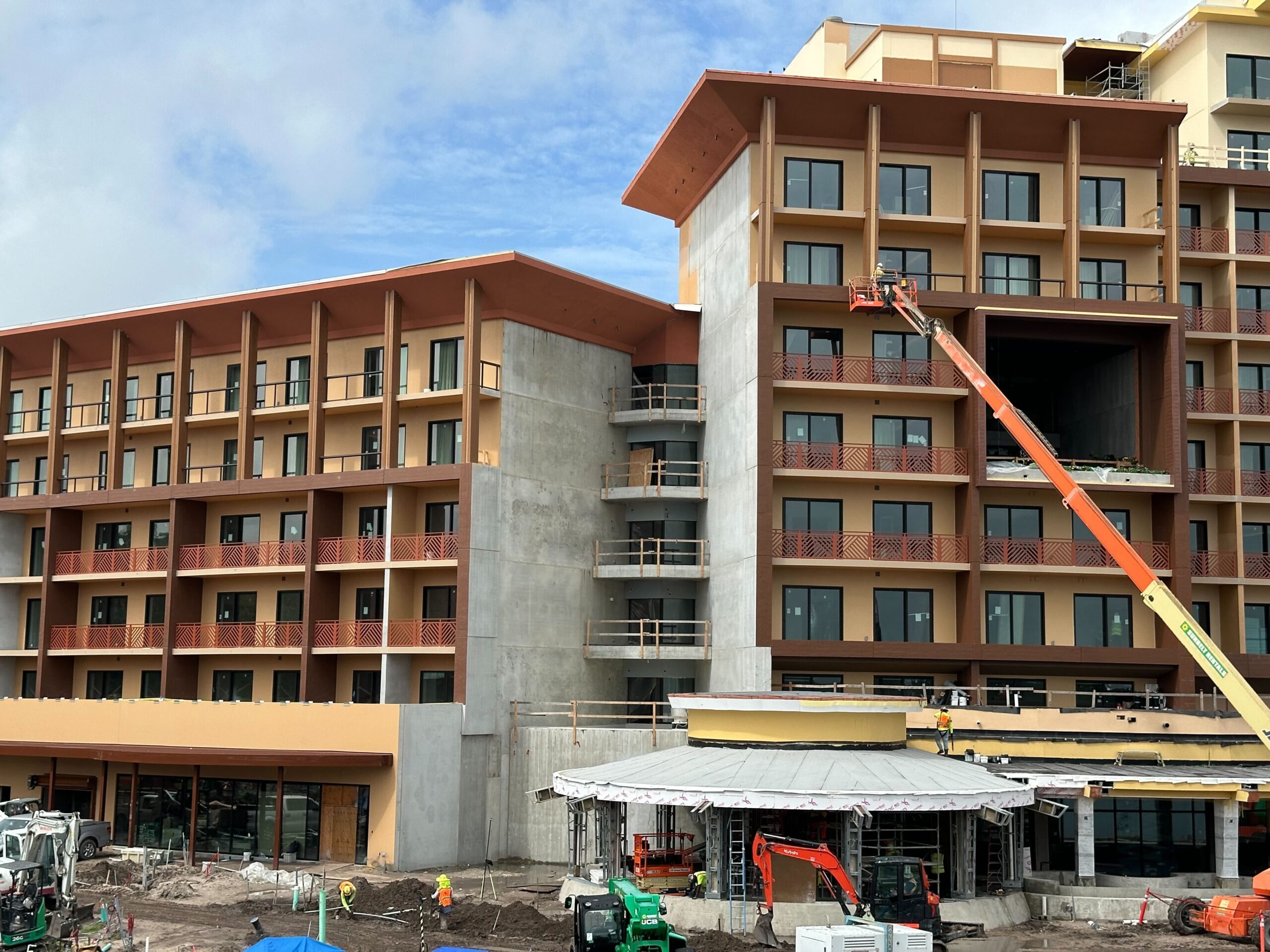 A mid-construction multi-story building with multiple windows and balconies. Construction equipment and workers are visible on the ground and on the building, with a clear blue sky above.