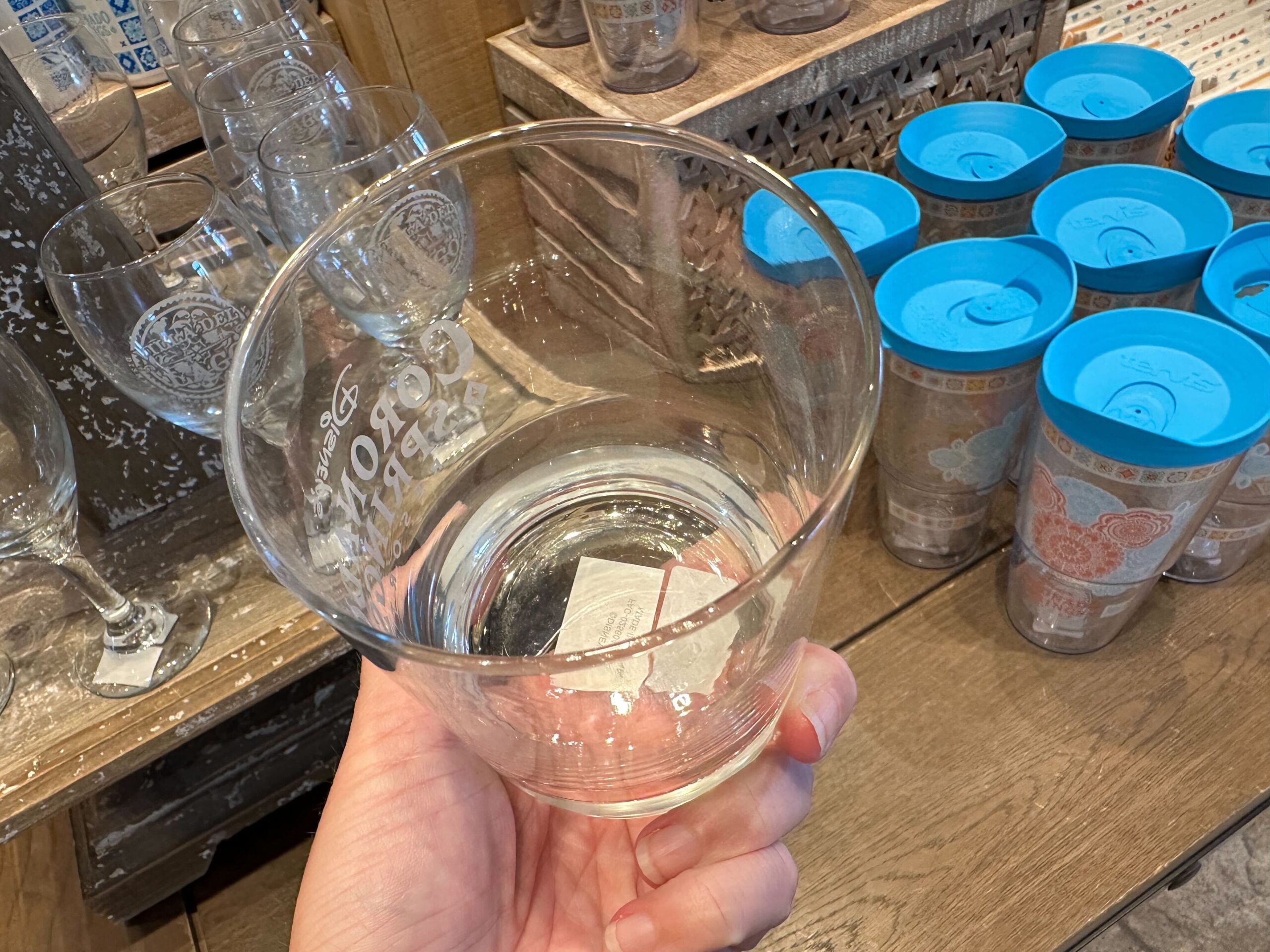 A hand holding an empty clear glass with a textured design, reminiscent of the intricate patterns found on Coronado Springs Merchandise, stands in front of shelves filled with glassware and blue-lidded containers.