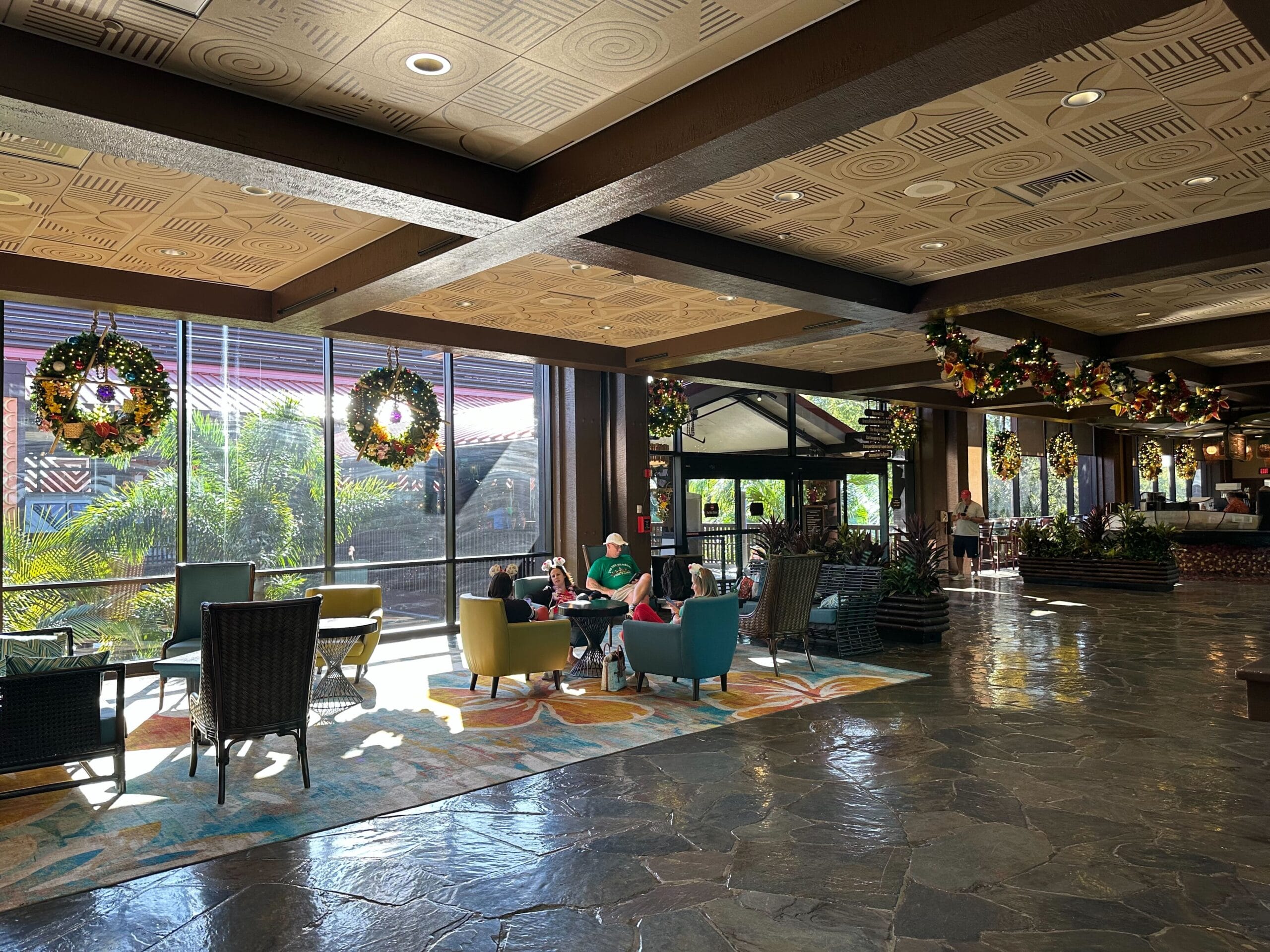 A hotel lobby with people seated in a sunlit area. Large windows reveal tropical plants outside. Wreaths hang from the ceiling, and chairs are arranged around tables on a patterned rug.