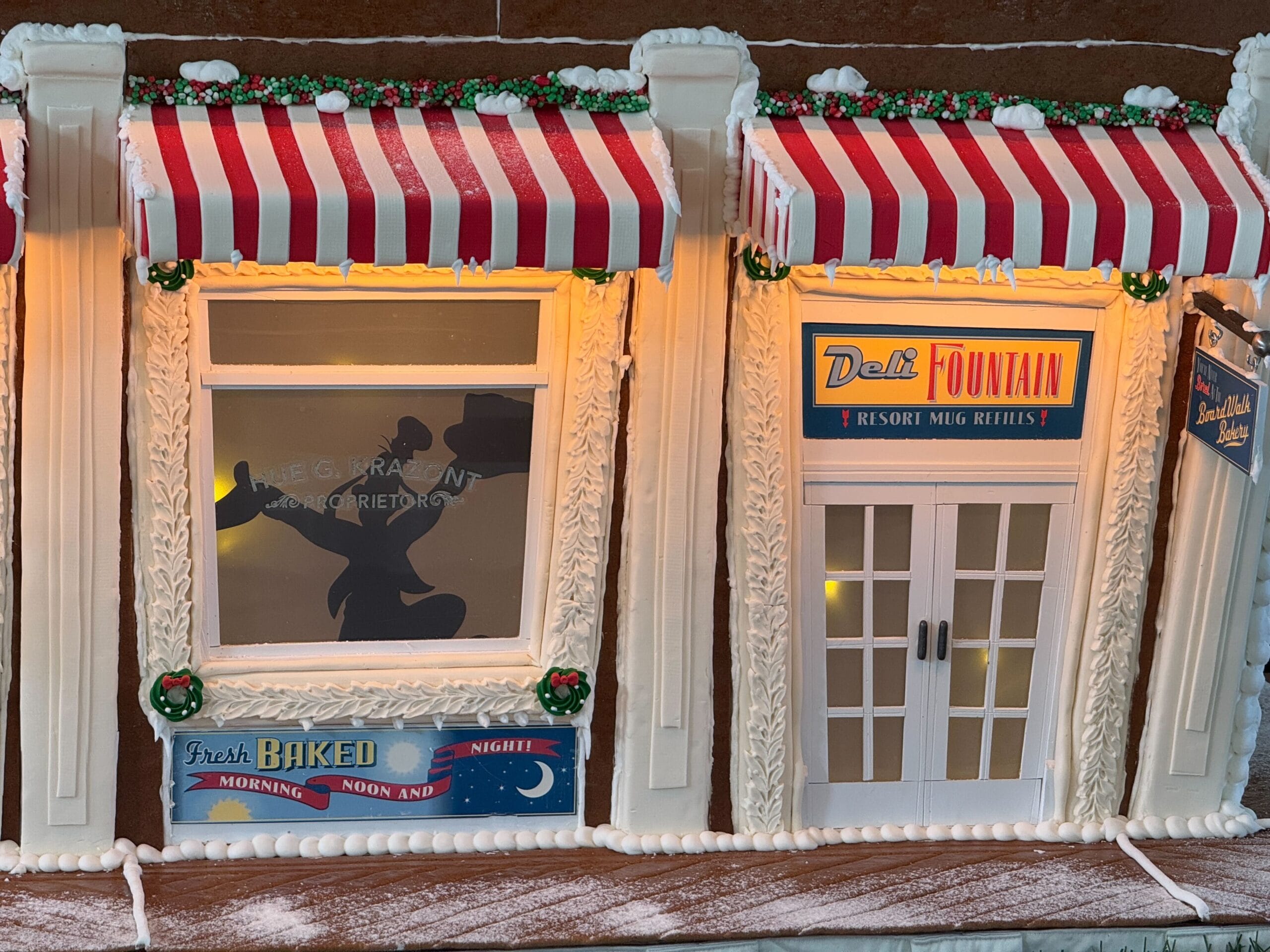 Gingerbread storefront with red and white striped awnings, illuminated windows, and festive wreaths, labeled Deli Fountain and Fresh Baked Morning Noon And Night.