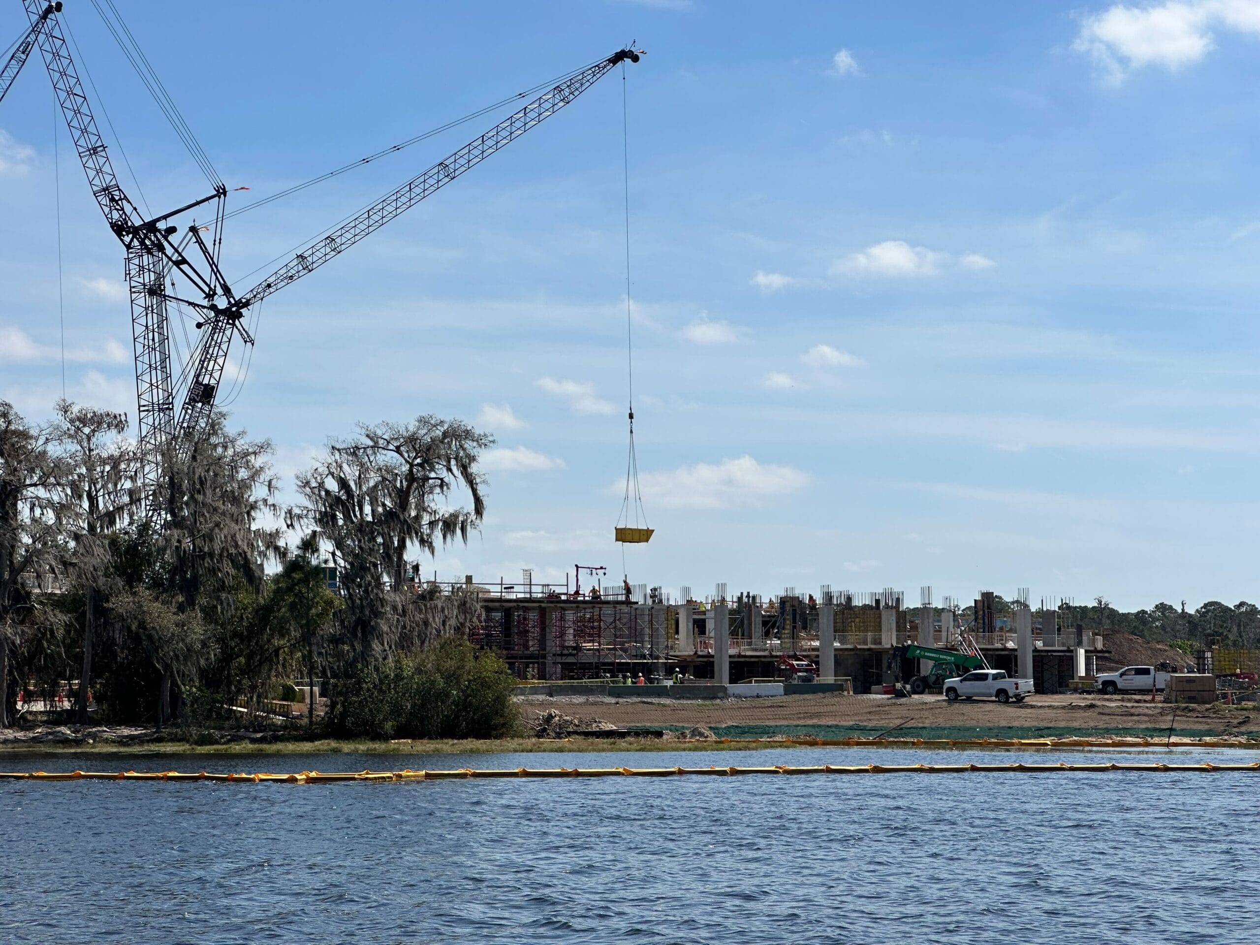 Construction update: The site near the water is bustling with activity as a crane lifts materials. Scaffolding and partially built structures rise against a backdrop of trees and clear sky, while access to the beach is temporarily blocked for safety.