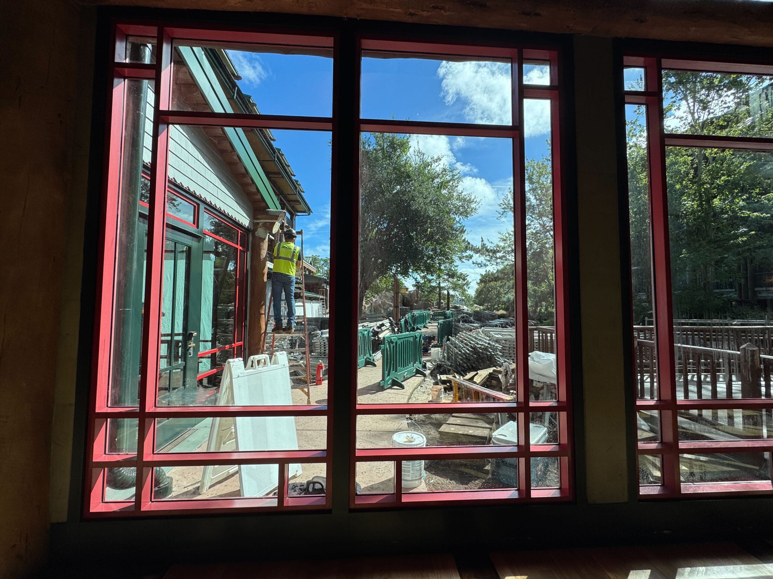 View through large windows of a worker on a ladder outside, surrounded by construction materials and greenery.