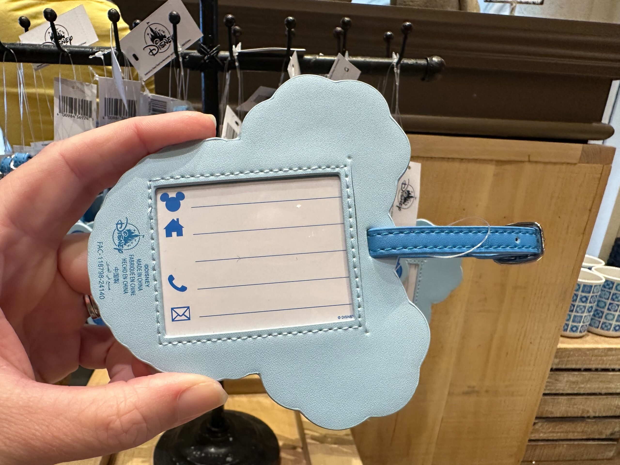 A hand holds a cloud-shaped Lug bag luggage tag with spaces for name, address, phone, and email. The tag is light blue with a leather strap, reminiscent of Coronado Springs merchandise in its unique design.