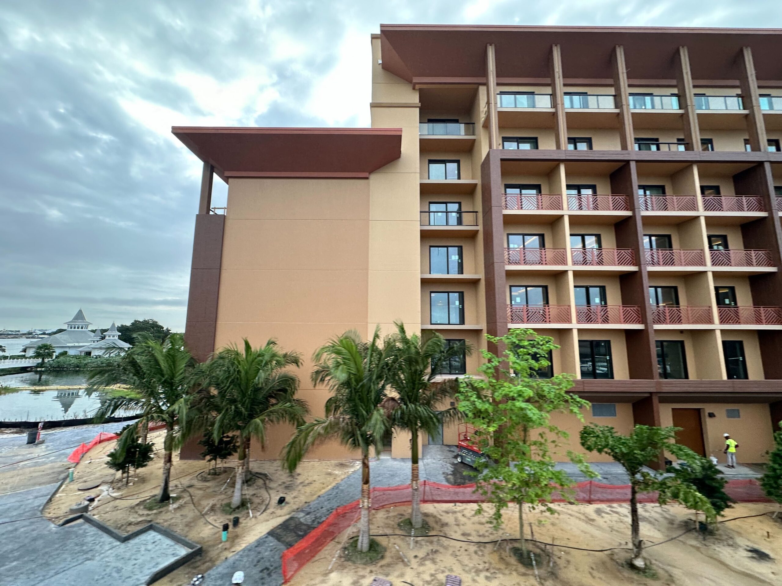 A modern multistory building with balconies under a cloudy sky. Palms trees and a partially completed pathway are in the foreground, with a worker visible near the trees.