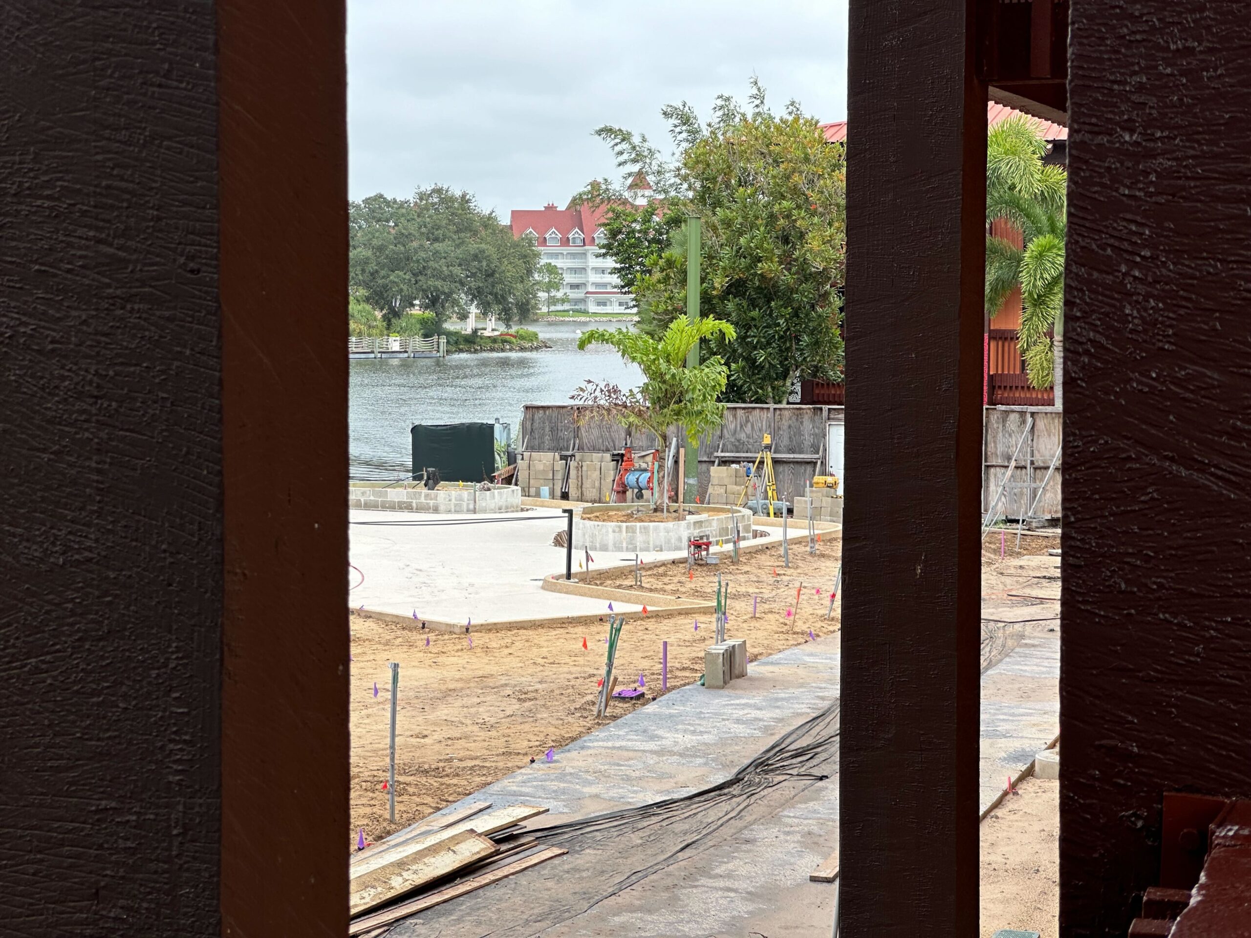 View of a construction site with concrete foundations, surrounded by trees and buildings near a body of water. Various construction materials and tools are scattered around the area.