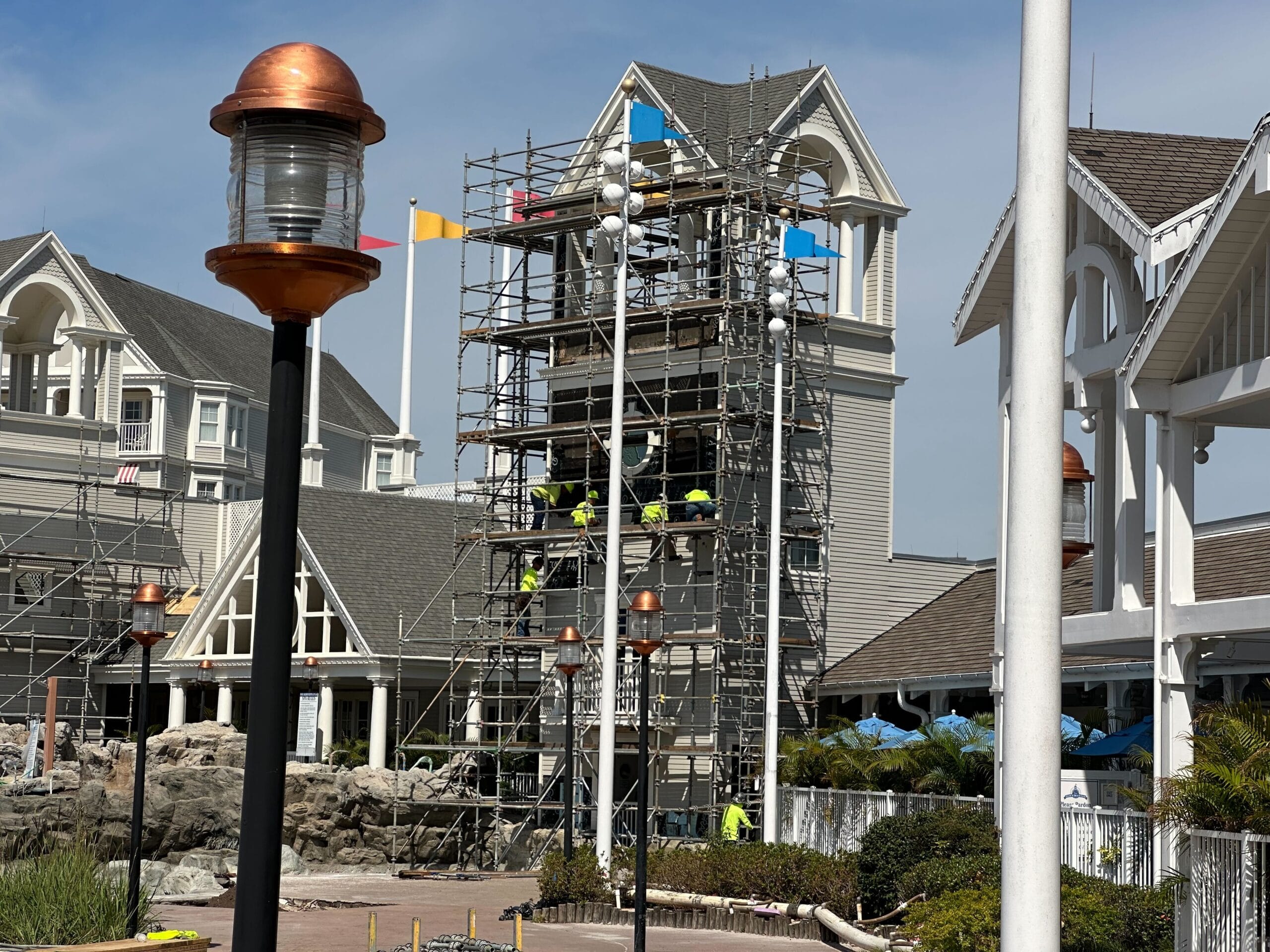 A building under renovation with a tower of new scaffolding and construction workers. Two adjacent structures boast gabled roofs. Elegant lamp posts stand in the foreground, while flags flutter on poles, reminiscent of Disney’s Yacht Club charm.