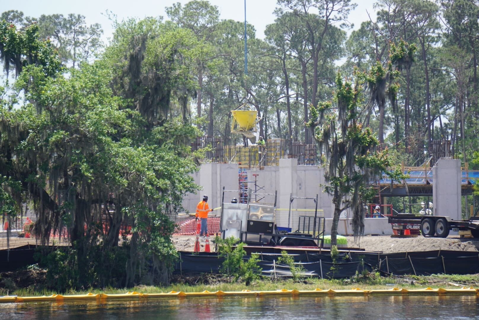 A construction site with workers and equipment near a riverbank. Concrete structures are being erected, and trees surround the area.