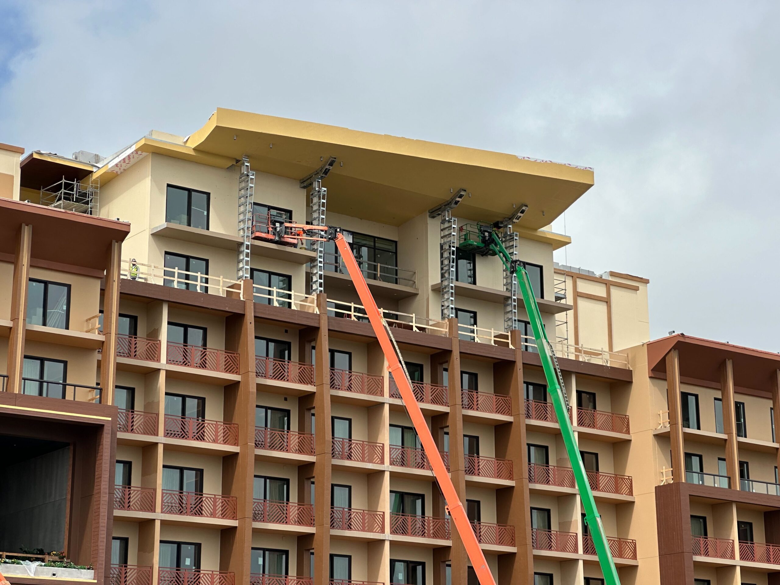 A construction site showing workers on cranes installing structural elements on the top floors of a multi-story building.
