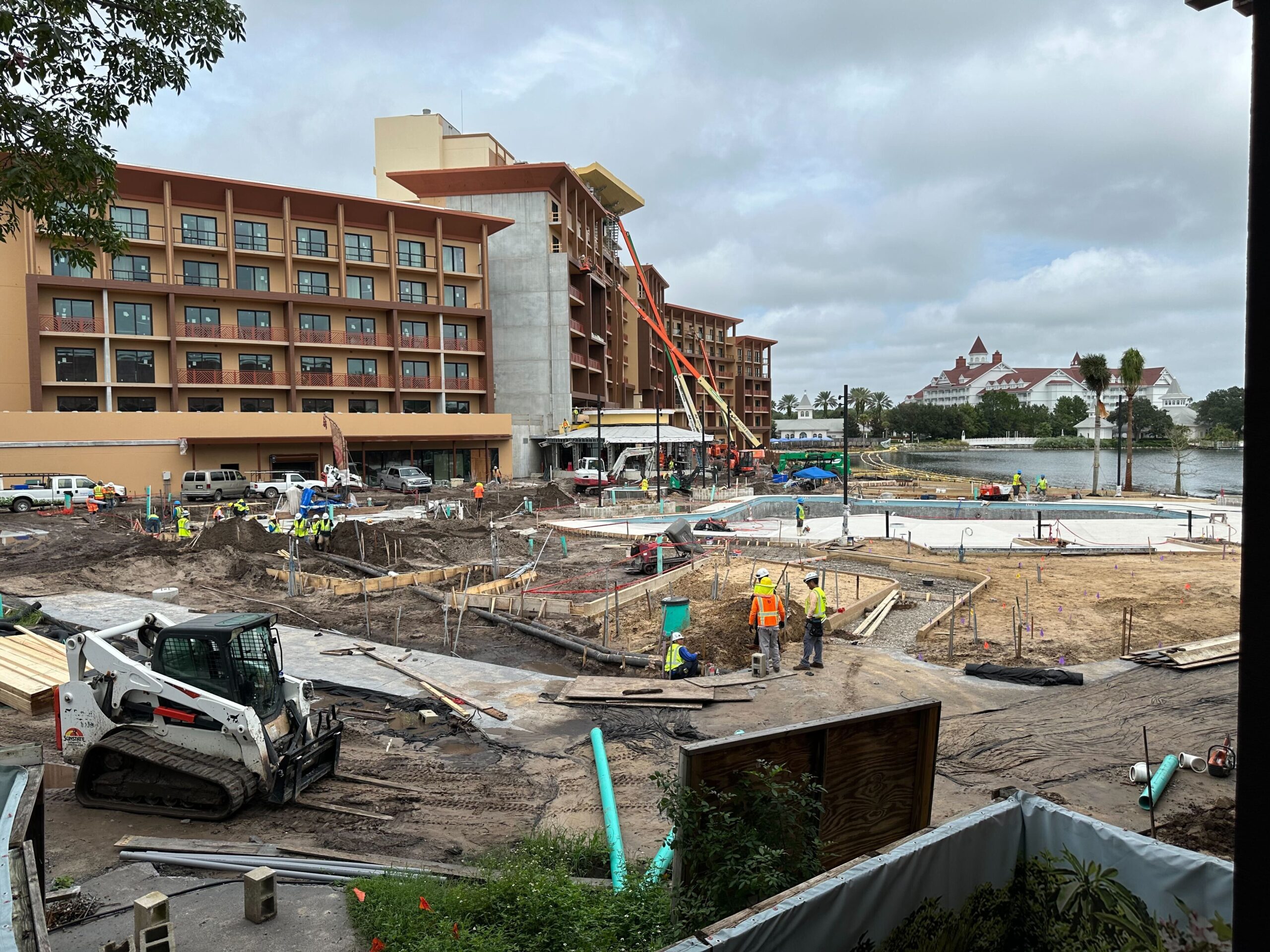 Construction site with workers and machinery in front of a multi-story hotel building. Scaffolding is set up along the building while the construction of outdoor areas is ongoing.