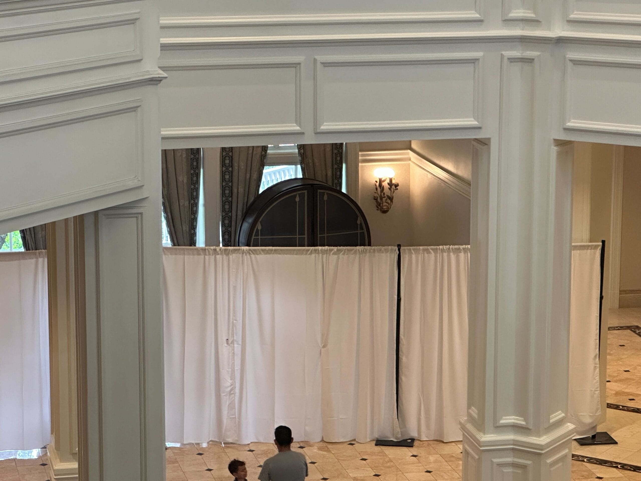Two people stand in a large lobby area with white curtains set up as partitions near a window and wall sconce.