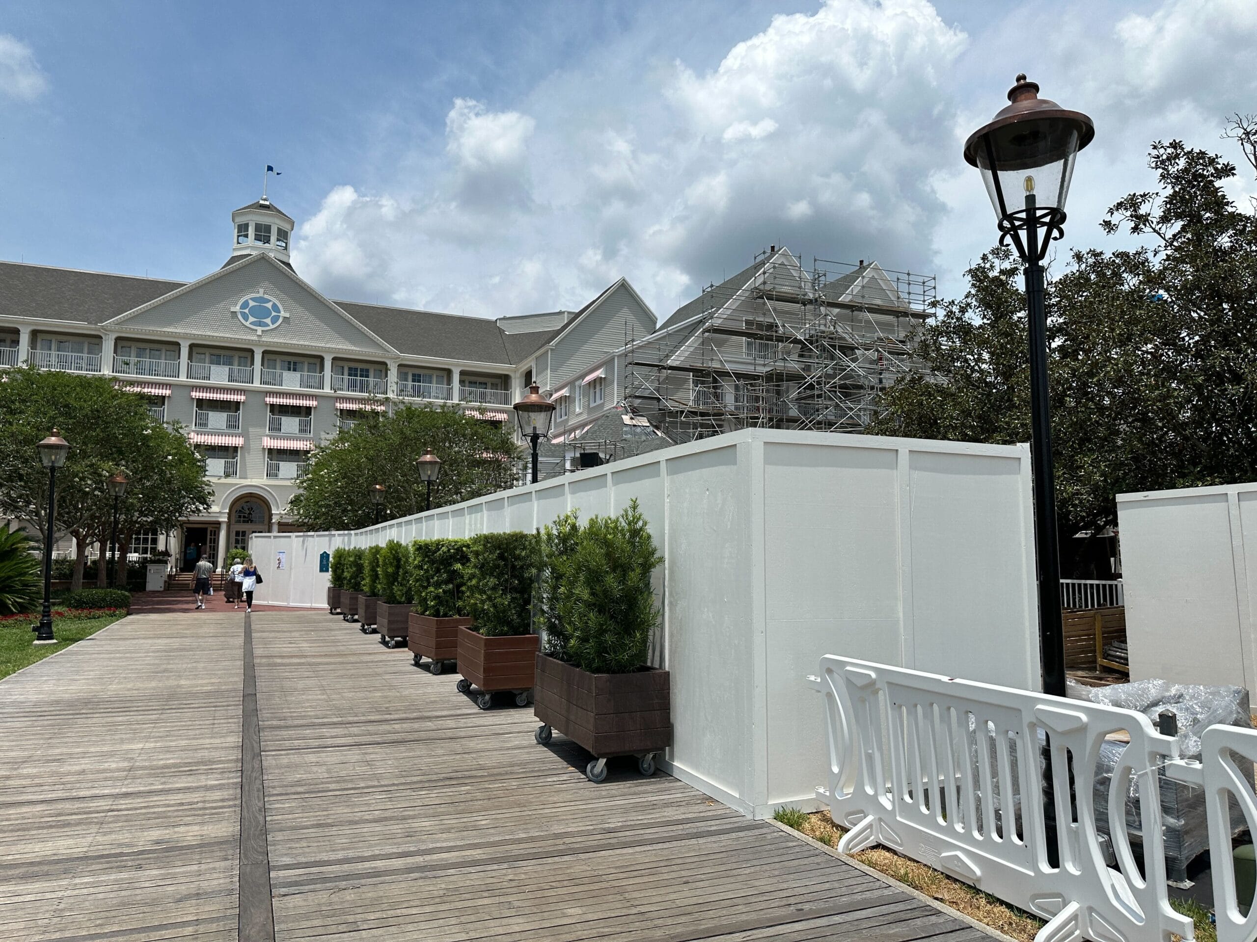 A hotel with construction scaffolding and white barriers along a wooden walkway lined with potted plants.