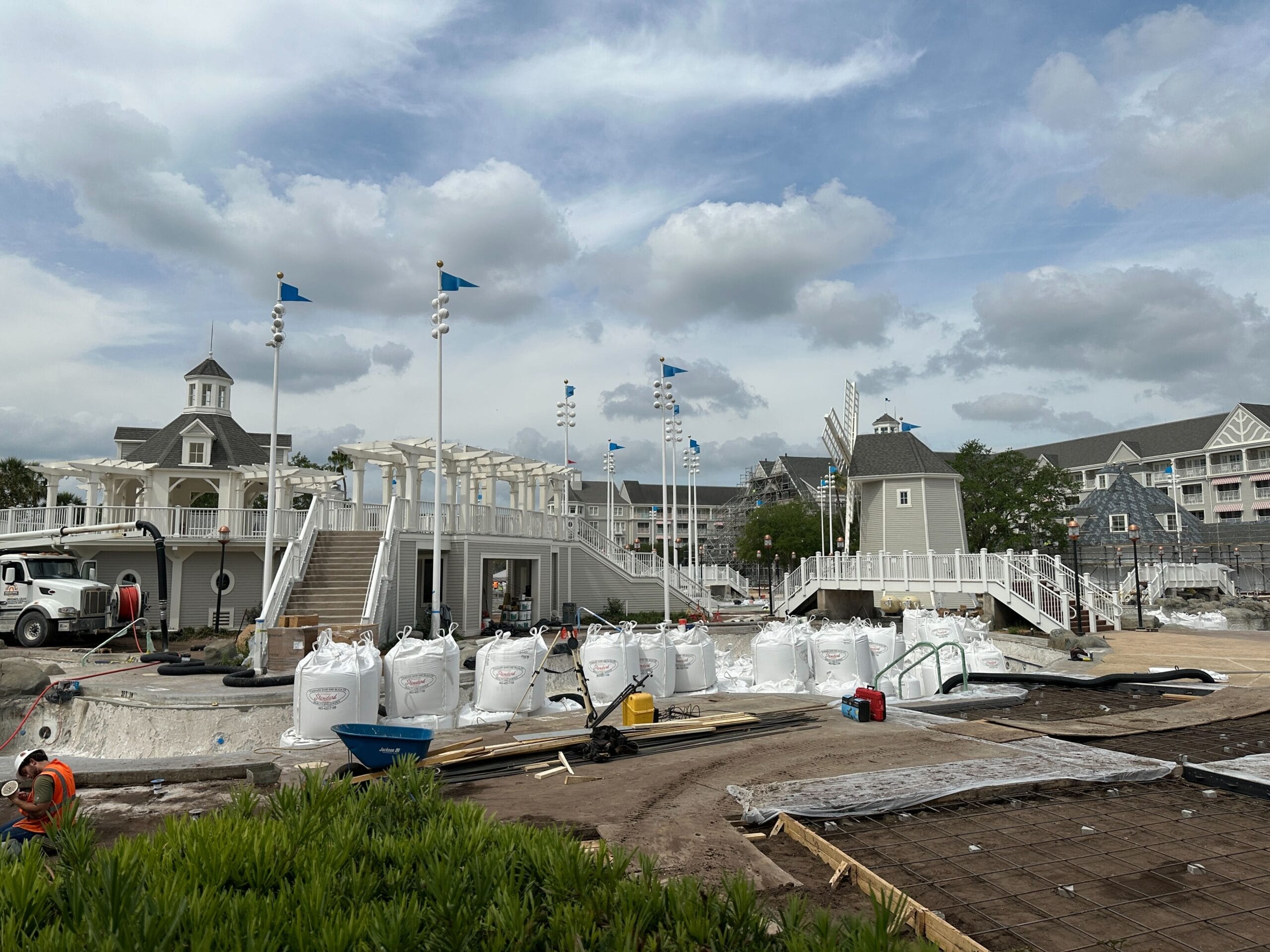 Construction site with white bags, tools, and fencing in front of a large, light gray Victorian-style building under a cloudy sky.