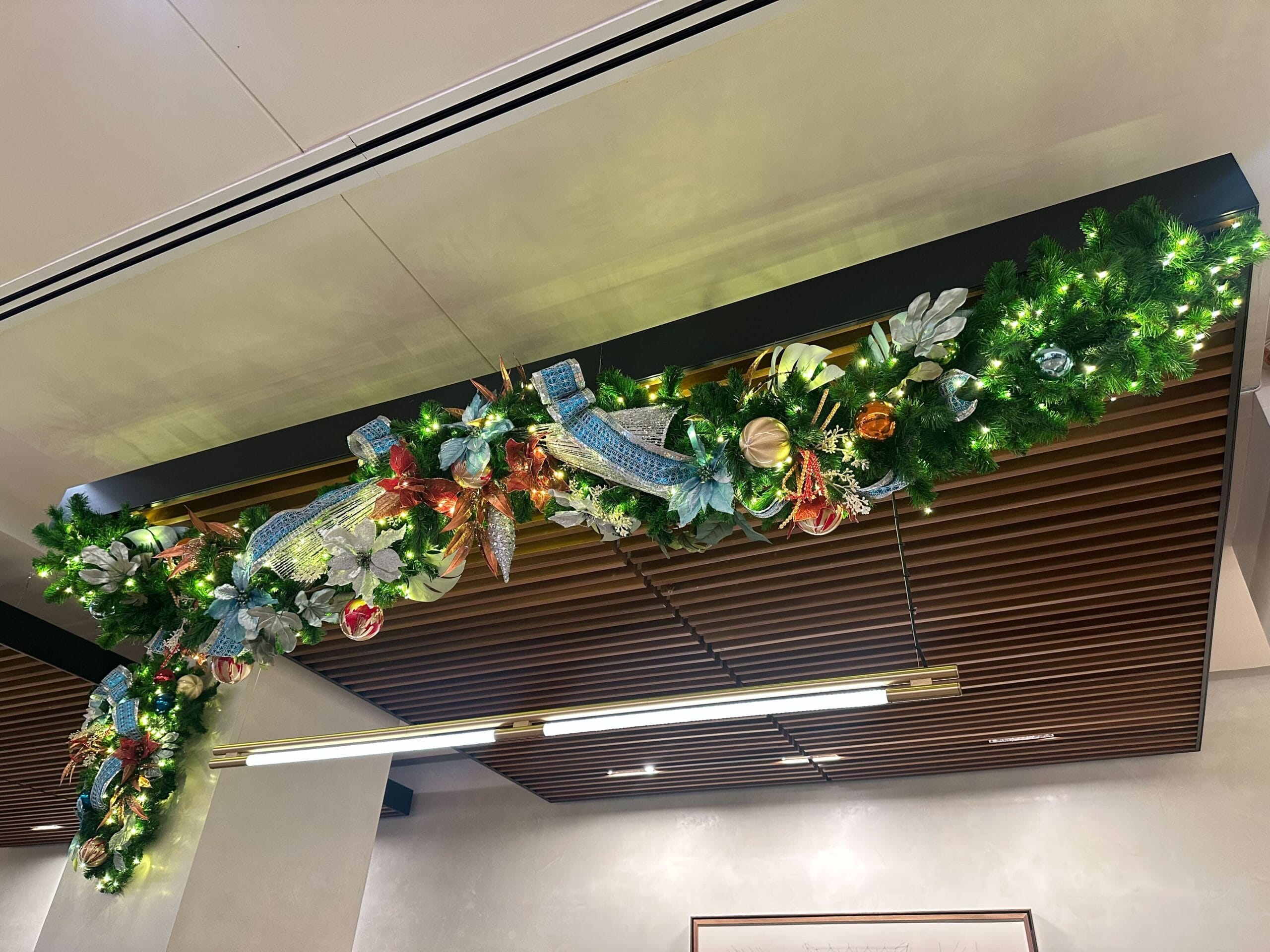 A festive garland with green foliage, colorful ornaments, and ribbon, decorated with lights, hangs from a wooden ceiling.