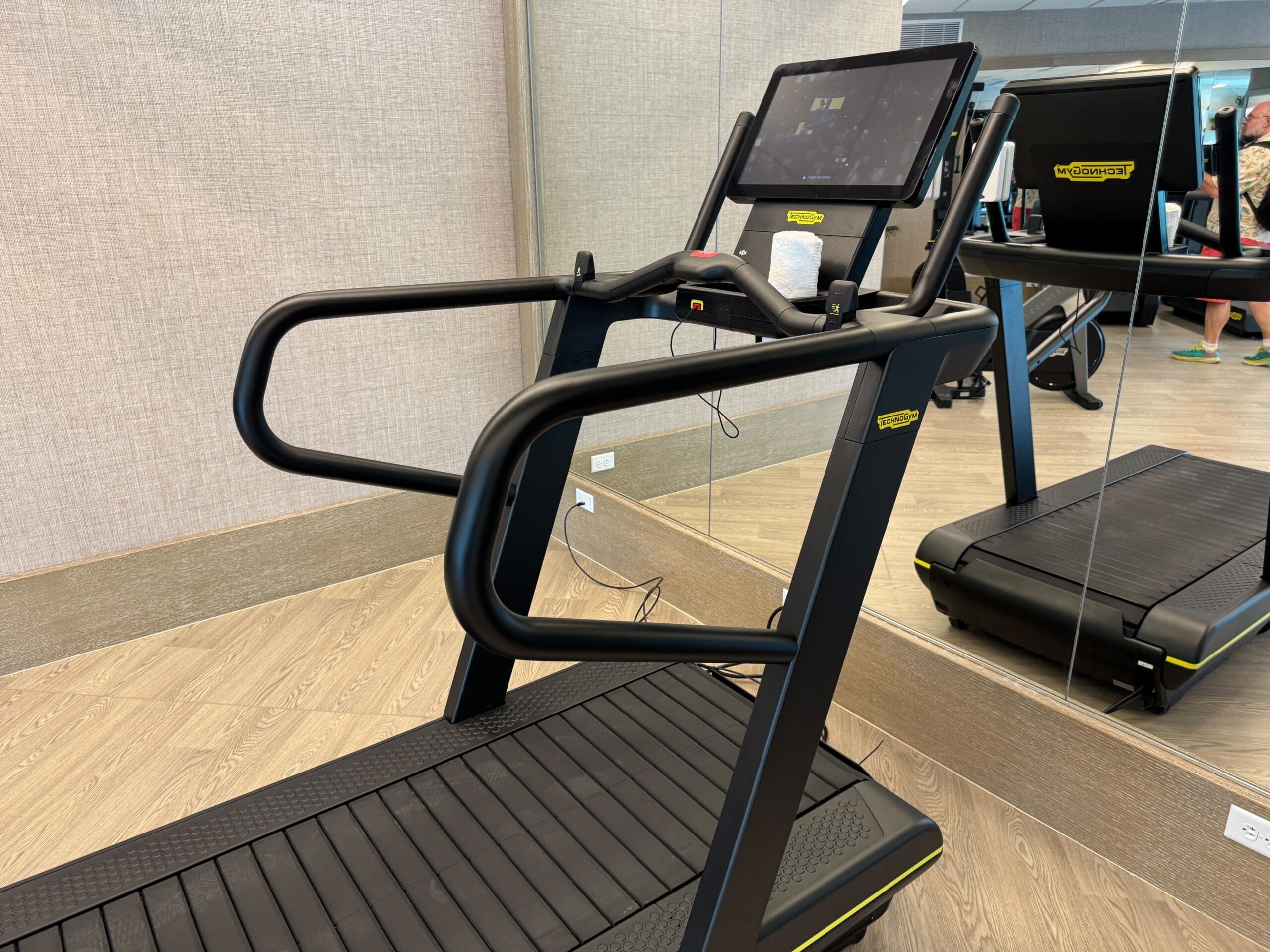 A treadmill with handrails and a tablet holder is positioned in a gym with a mirror and wooden floor.