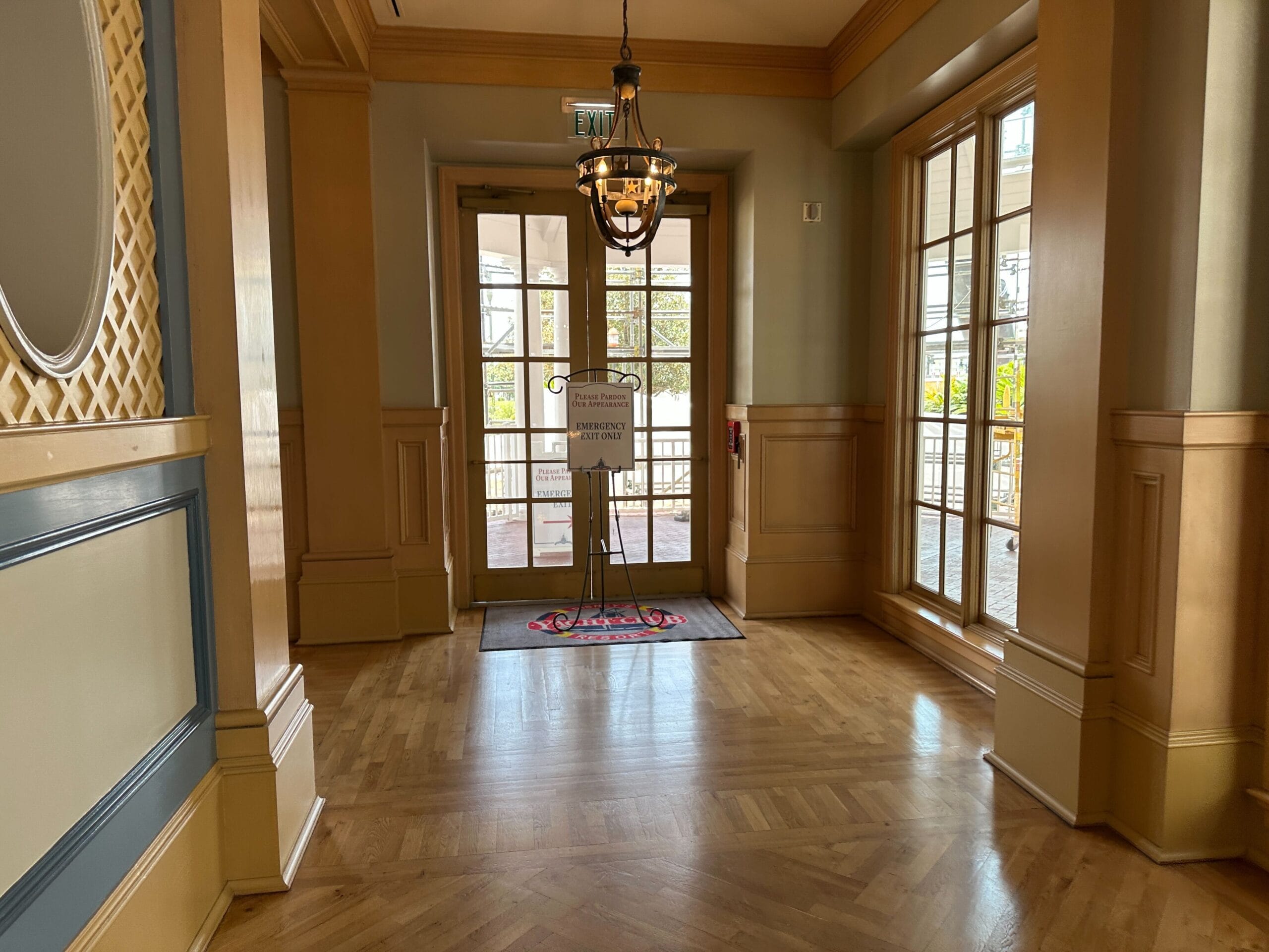 A bright hallway with wood floors, large windows, and a glass door leading outside with a sign in front.