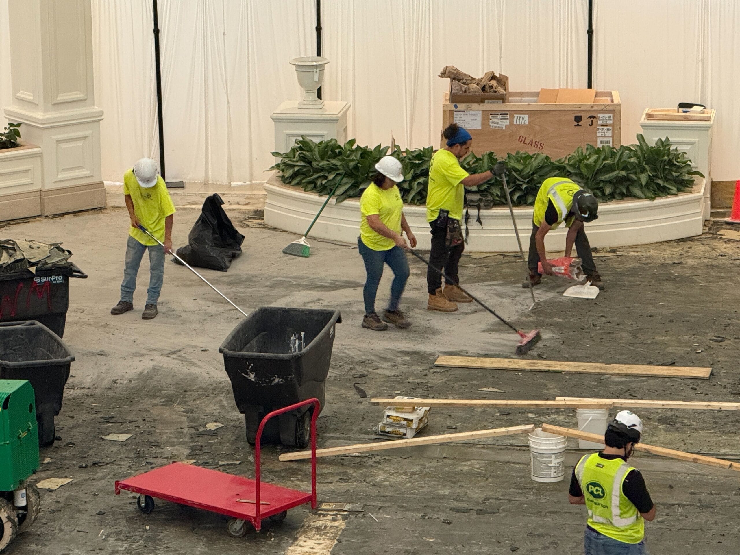 Five construction workers in yellow vests clean debris from a large indoor area with brooms and shovels.