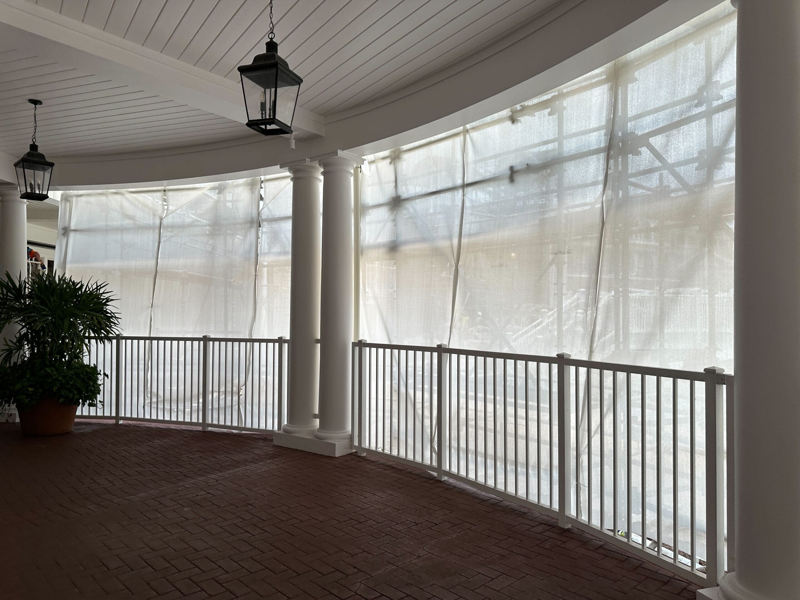 Covered porch with white columns and railings, brick floor, hanging lanterns, and sheer curtains letting in filtered light.