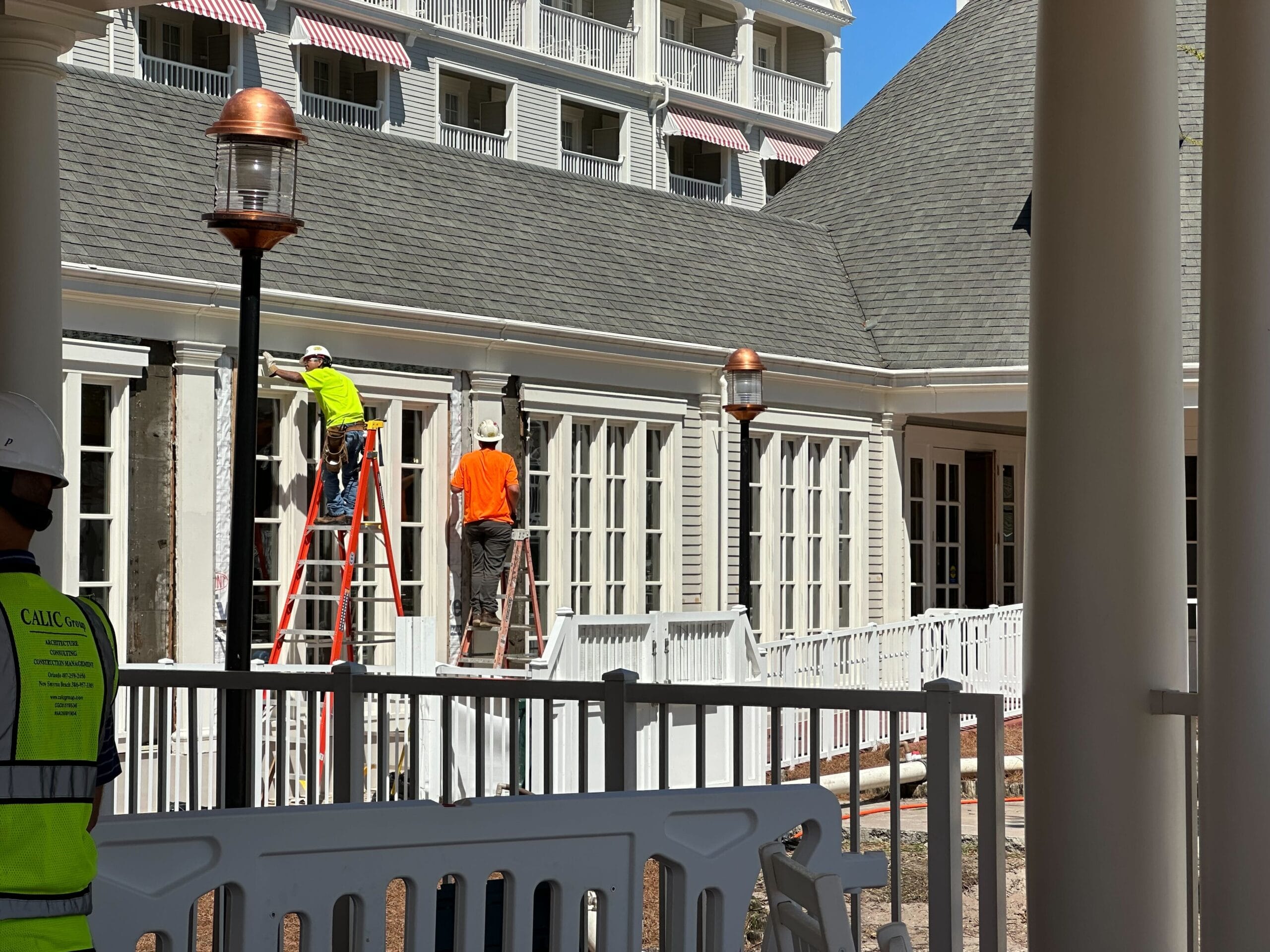 Construction workers on ladders repairing a building exterior with white walls and large windows, under a clear blue sky.