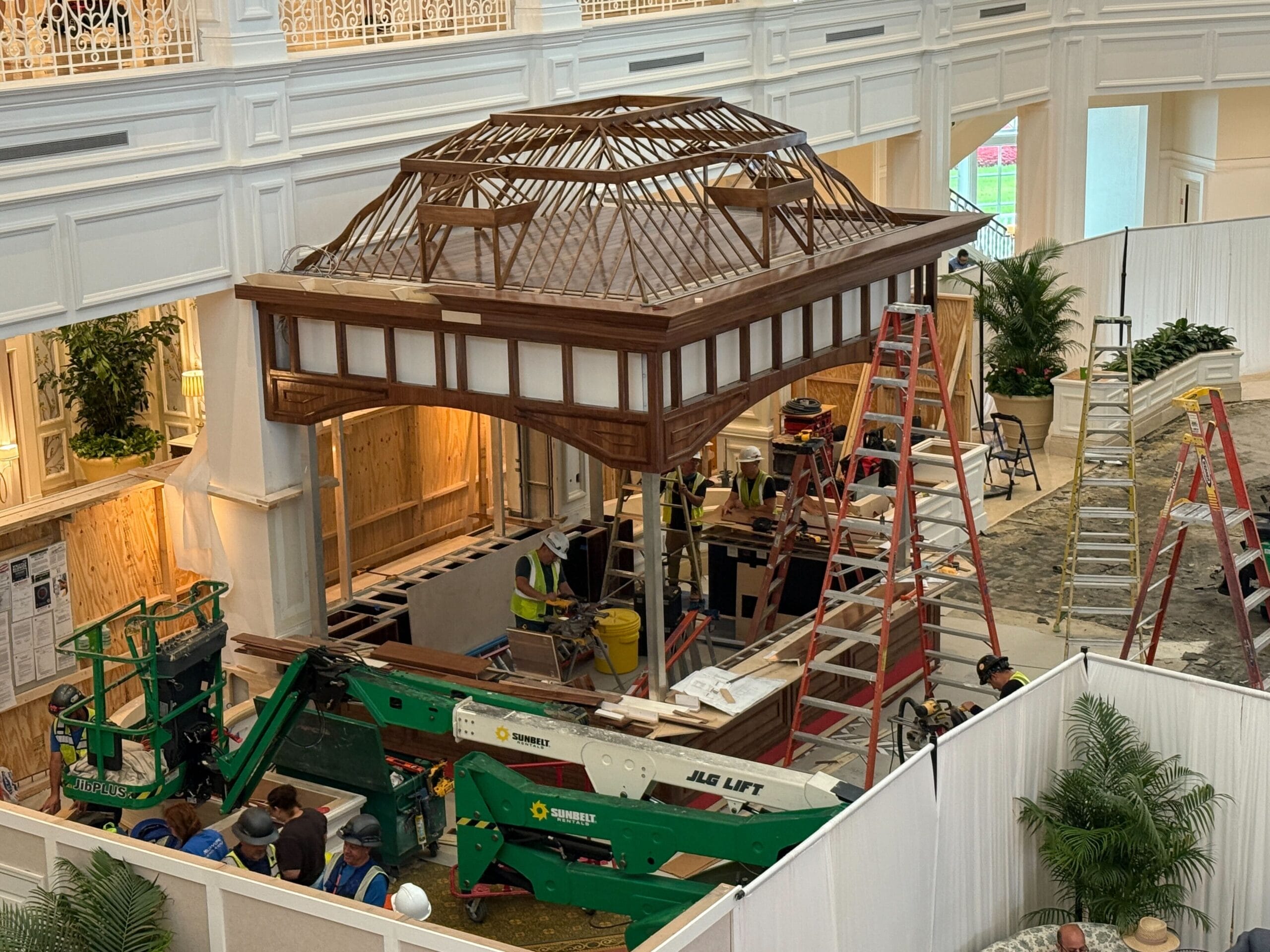 Construction workers assemble a large wooden gazebo structure indoors, surrounded by ladders and construction equipment.