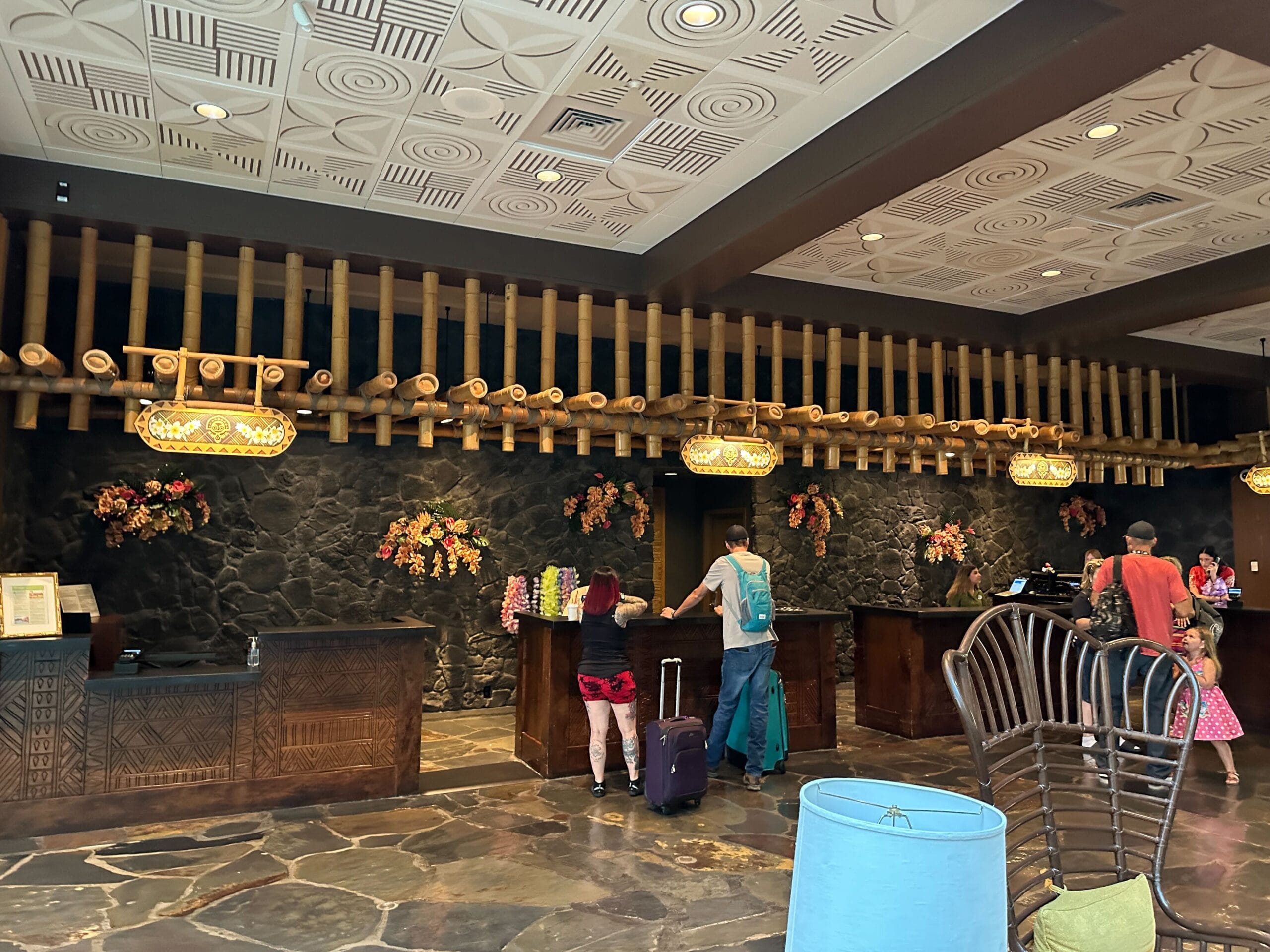 Hotel front desk with guests checking in, floral decor, suitcases, and blocked off lobby seating at Disney's Polynesian Village Resort.