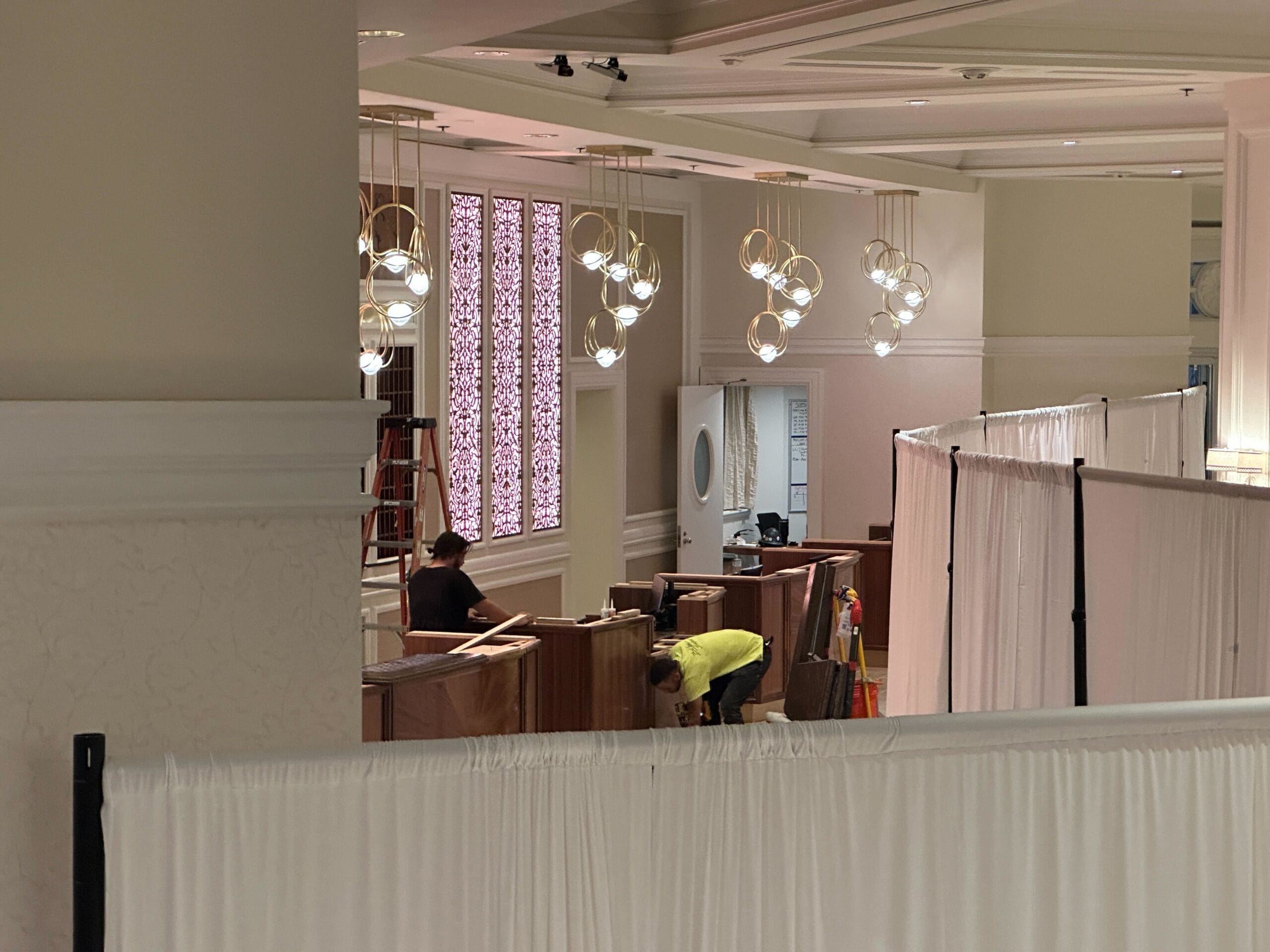 Two people set up wooden booths behind white curtains in Disney's Grand Floridian with hanging lights and decorative wall panels.
