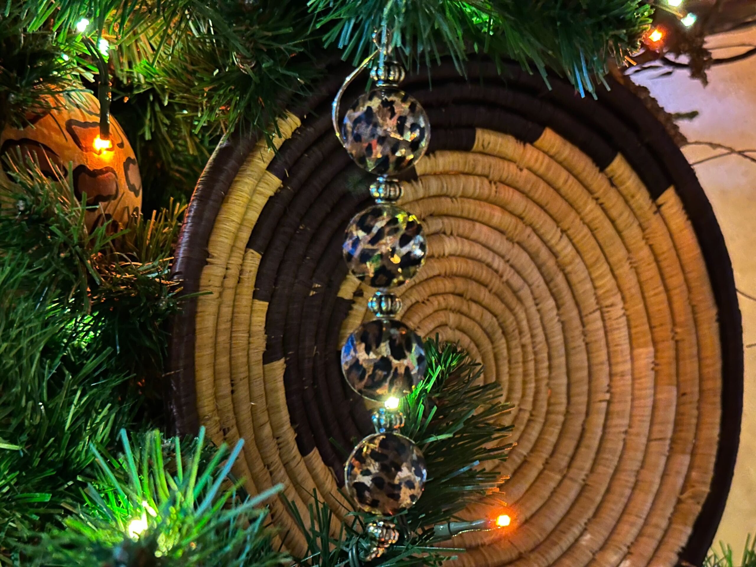 Beaded ornament with animal print hangs on a Christmas tree, surrounded by green branches and colorful lights. A woven circular design is visible in the background.
