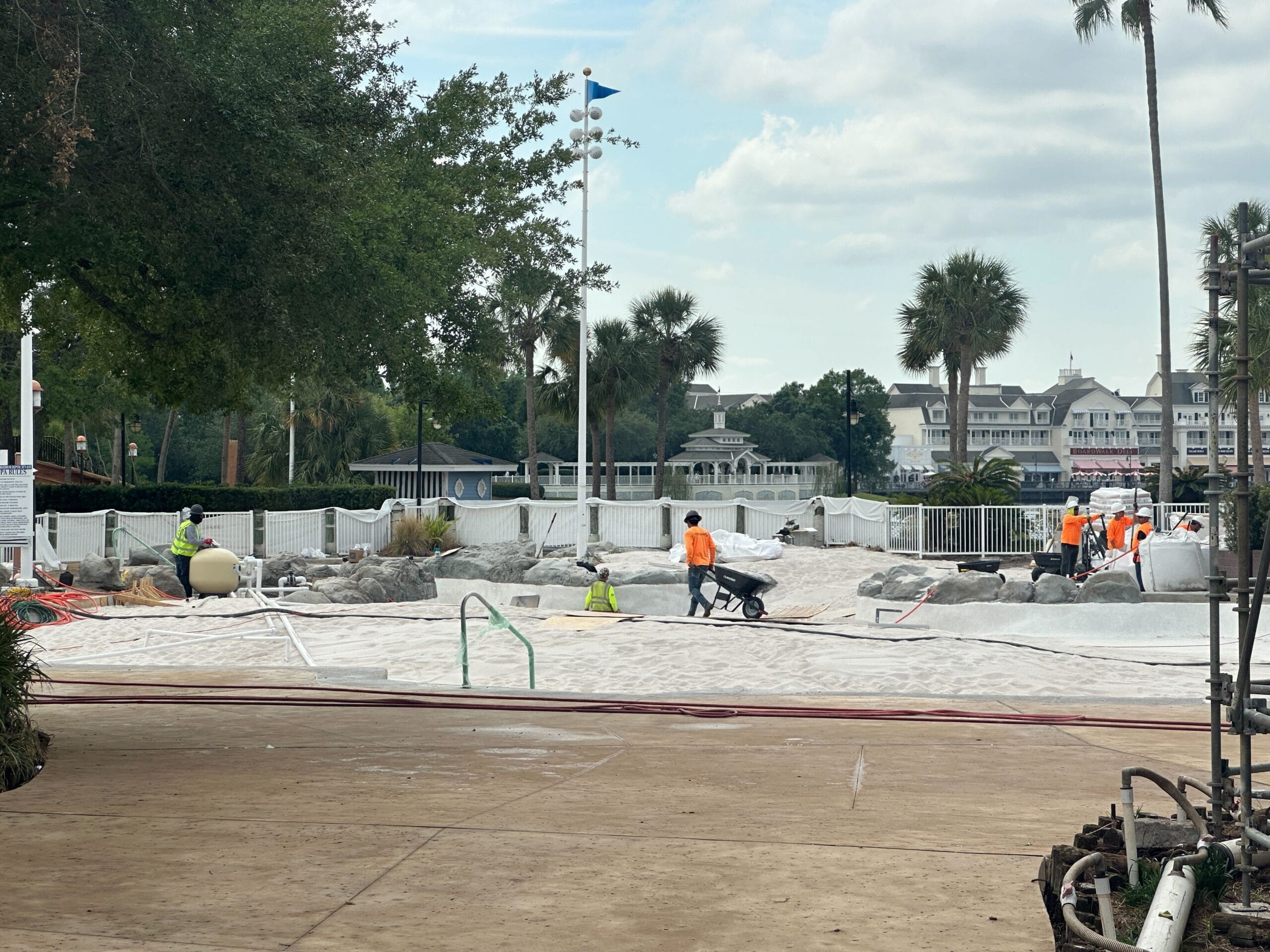 Construction workers in orange vests work on a sandy area near a pool, with palm trees and buildings in the background.