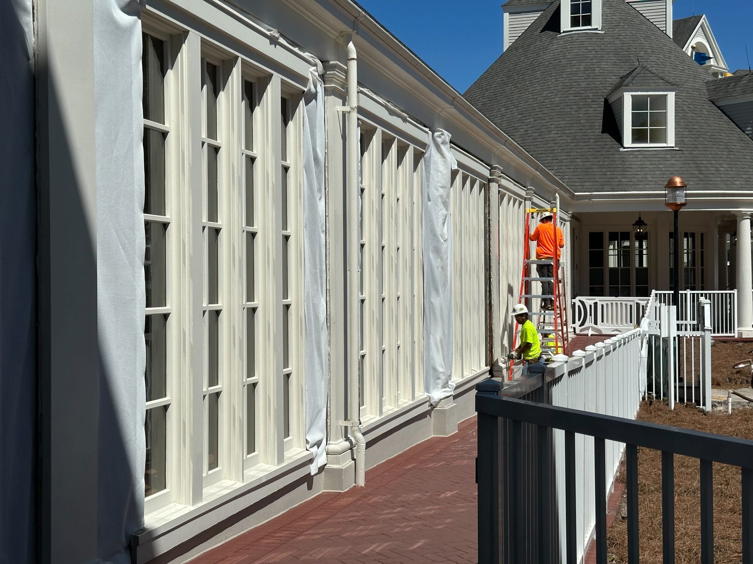 Workers in safety vests are on ladders, painting or renovating the exterior of a white building with large windows and a sloped roof.
